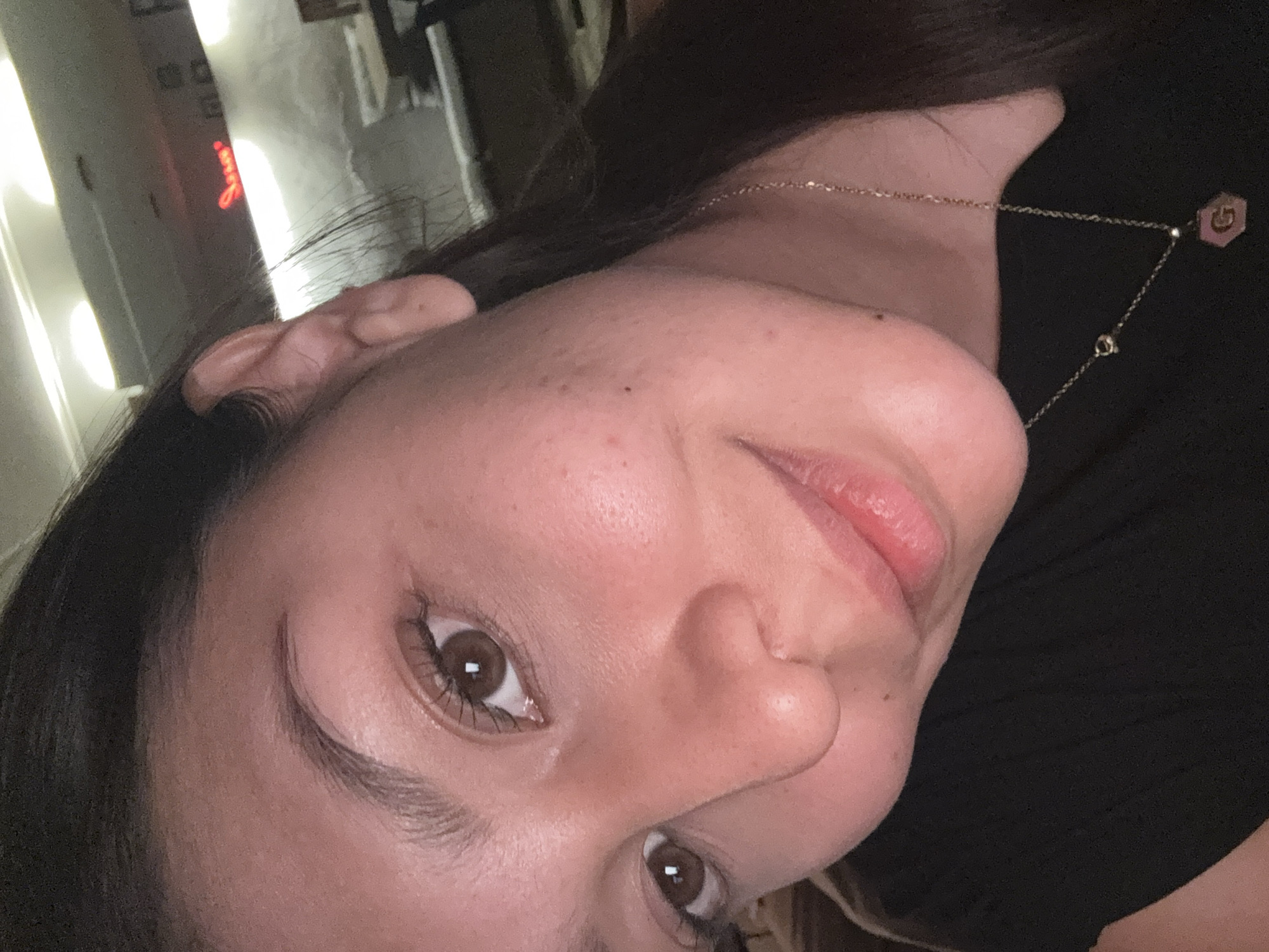 A close-up, tilted selfie of a woman with light skin, brown hair, and brown eyes. She is wearing a gold necklace with a hexagonal pendant. The background appears to be indoors, with blurred lighting and objects that are not identifiable. The location in San Andrés Cholula, Mexico, cannot be determined.
