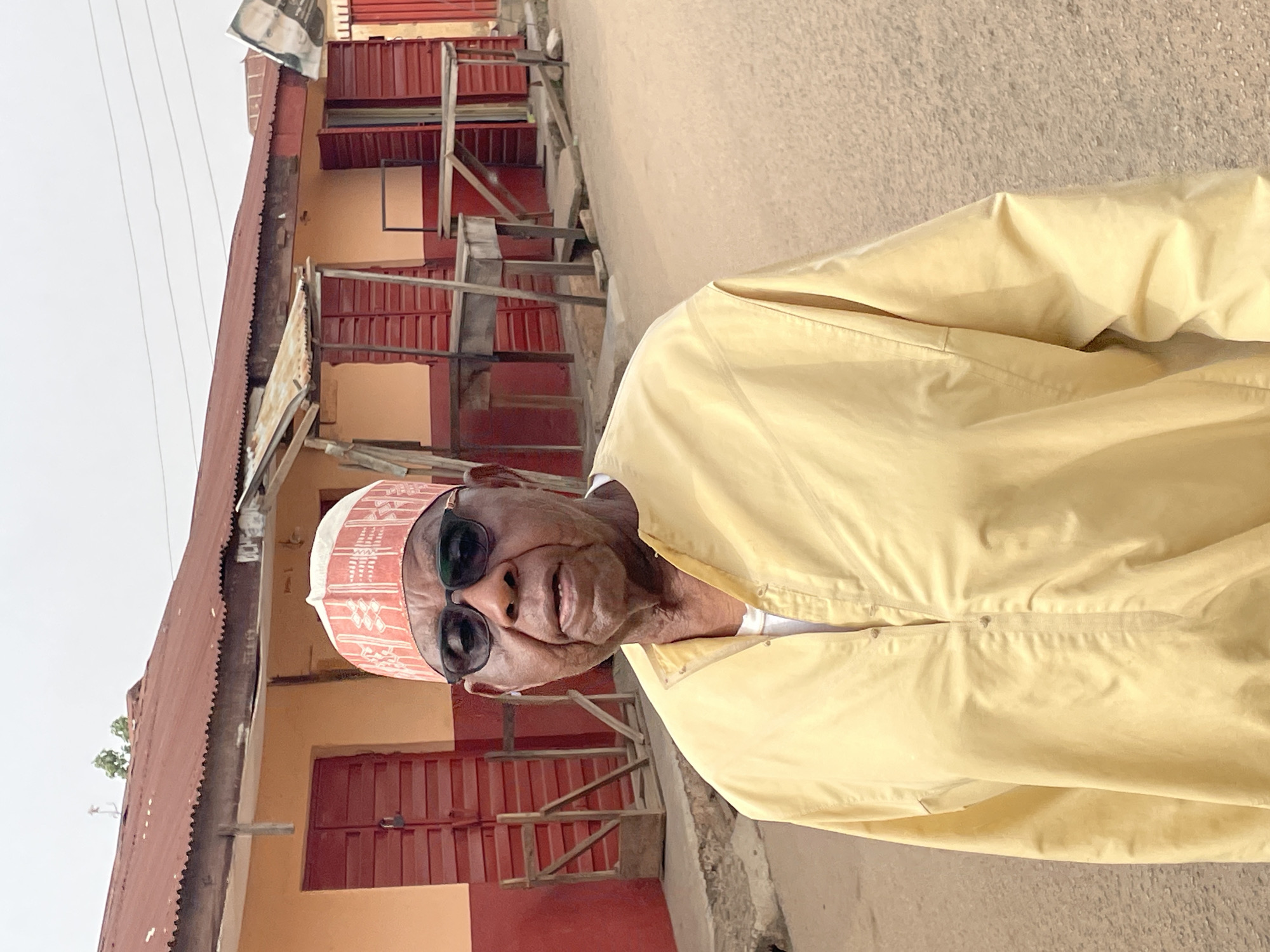 An elderly man is standing in front of a building with red doors in Jos, Nigeria. The man is wearing a light yellow garment, a white cap with red patterns, and sunglasses. He appears to be on a sidewalk. The building behind him has a weathered, red-painted roof. There is a makeshift wooden structure leaning against the building. The sky is overcast.