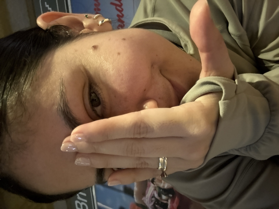 A woman with dark hair and a fair complexion is partially visible, with one eye open and looking directly at the viewer. Her hand, adorned with a silver ring, covers part of her face. She's wearing a light green hooded jacket. Her ear is visible, featuring multiple earrings. The background includes a sign with lettering. The image is rotated, presenting a tilted perspective.