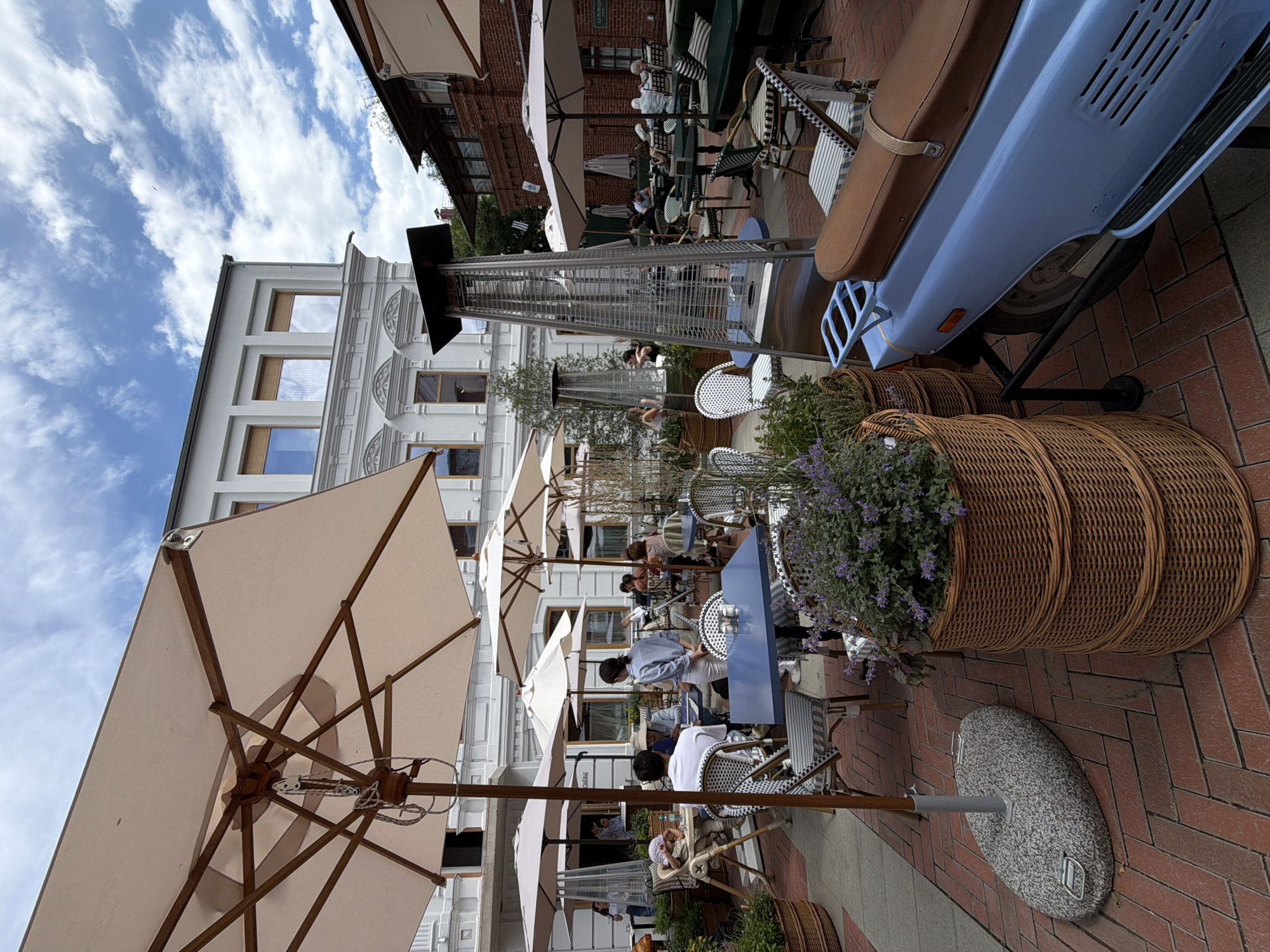 The image depicts an outdoor seating area of a cafe or restaurant, likely in Ufa, Russia. The scene is filled with tables and chairs set up on a brick-paved walkway. Several large, light-colored umbrellas provide shade over the seating. In the background, a white building with multiple windows and ornate architectural details is visible. A light blue scooter is parked on the right side of the image, with a brown leather seat. Large wicker baskets filled with plants and flowers are also placed around the seating area.
Several people are seated at the tables, some appearing to be patrons and others possibly staff. One person, possibly a waiter, is interacting with a table. Another person is pushing a stroller. The atmosphere seems relaxed and leisurely, suggesting it is daytime, likely late morning or afternoon, given the bright lighting and clear sky with some clouds. There are no overt signs of a specific event, but rather a typical day at an outdoor dining establishment.
One of the umbrellas shows a small black tag attached to its mechanism. The scooter has a red reflector and a visible license plate that is unreadable due to the resolution. A tall outdoor heater is positioned near the tables. The general ambiance is one of a pleasant, possibly warm, day, inviting people to dine outdoors.