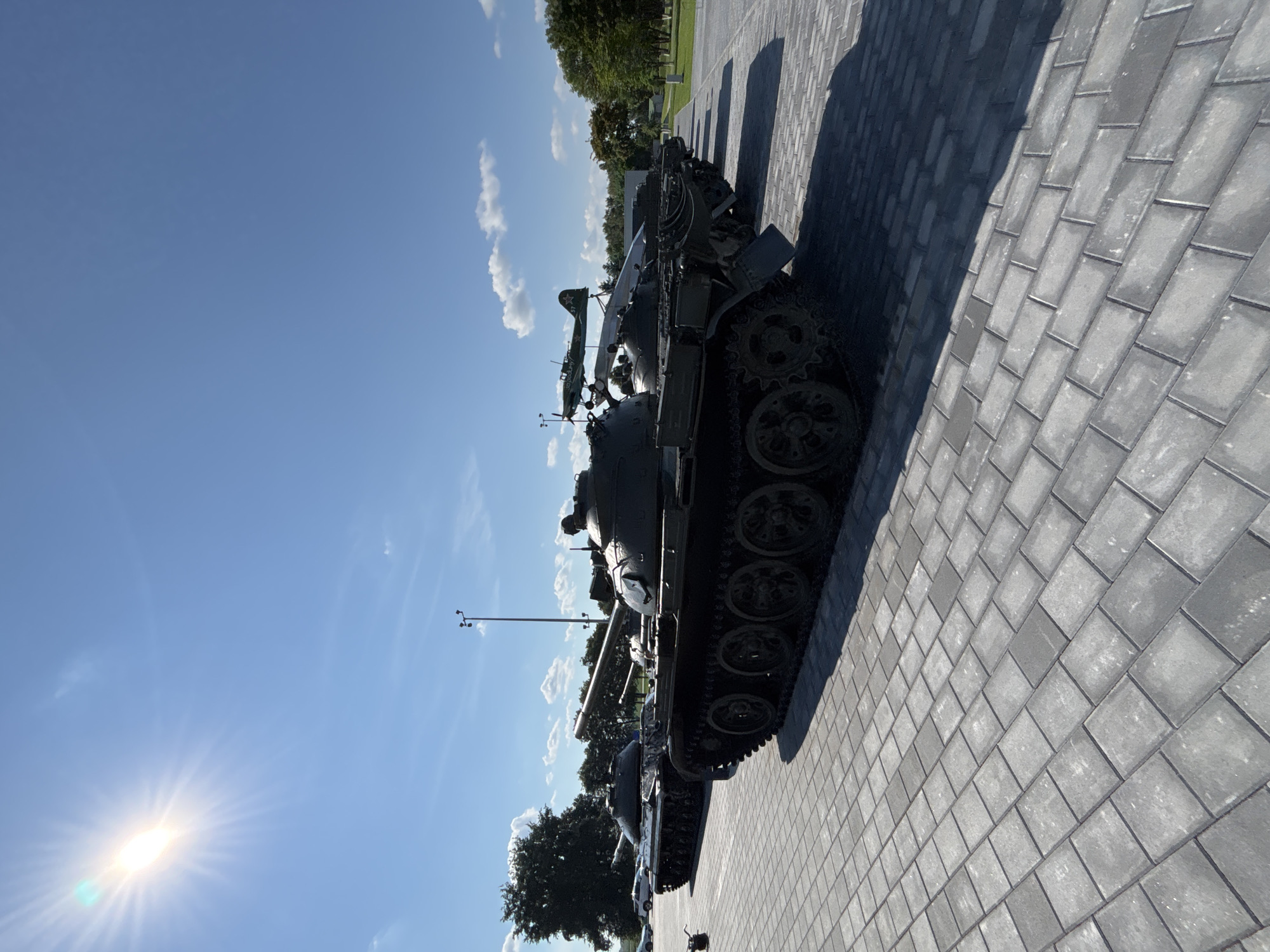 The image displays a row of several military tanks parked on a paved area with a cobblestone pattern. In the foreground, the tracks and wheels of the closest tank are prominent, casting a shadow onto the pavement. The tanks appear to be on display, possibly in a museum or memorial site. The background features a clear blue sky with the sun shining brightly in the upper left corner, creating a lens flare effect. Scattered white clouds are visible. Beyond the tanks, trees and greenery line the horizon, and a lamppost stands tall. A small propeller airplane is mounted on a structure behind one of the tanks. The location is identified as the city of Ozyaritska-Slabadski rural council, Belarus. There are no people visible in the image, and the tanks are stationary.