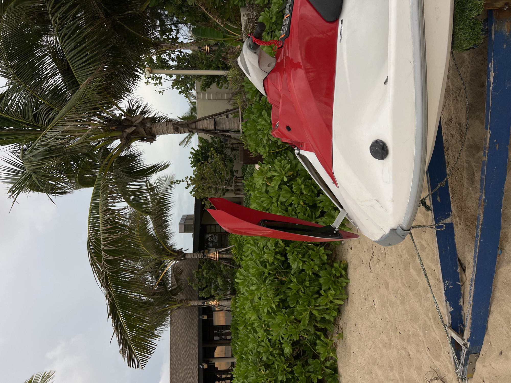 A red and white jet ski is parked on a sandy beach, resting on a blue trailer. The trailer is partially submerged in the sand. Behind the jet ski, a dense green hedge lines the edge of the beach. Further back, a low-rise building with a brown roof and large windows is visible, with several palm trees in front of it. The sky is overcast. The scene is quiet and appears to be a resort or private property.