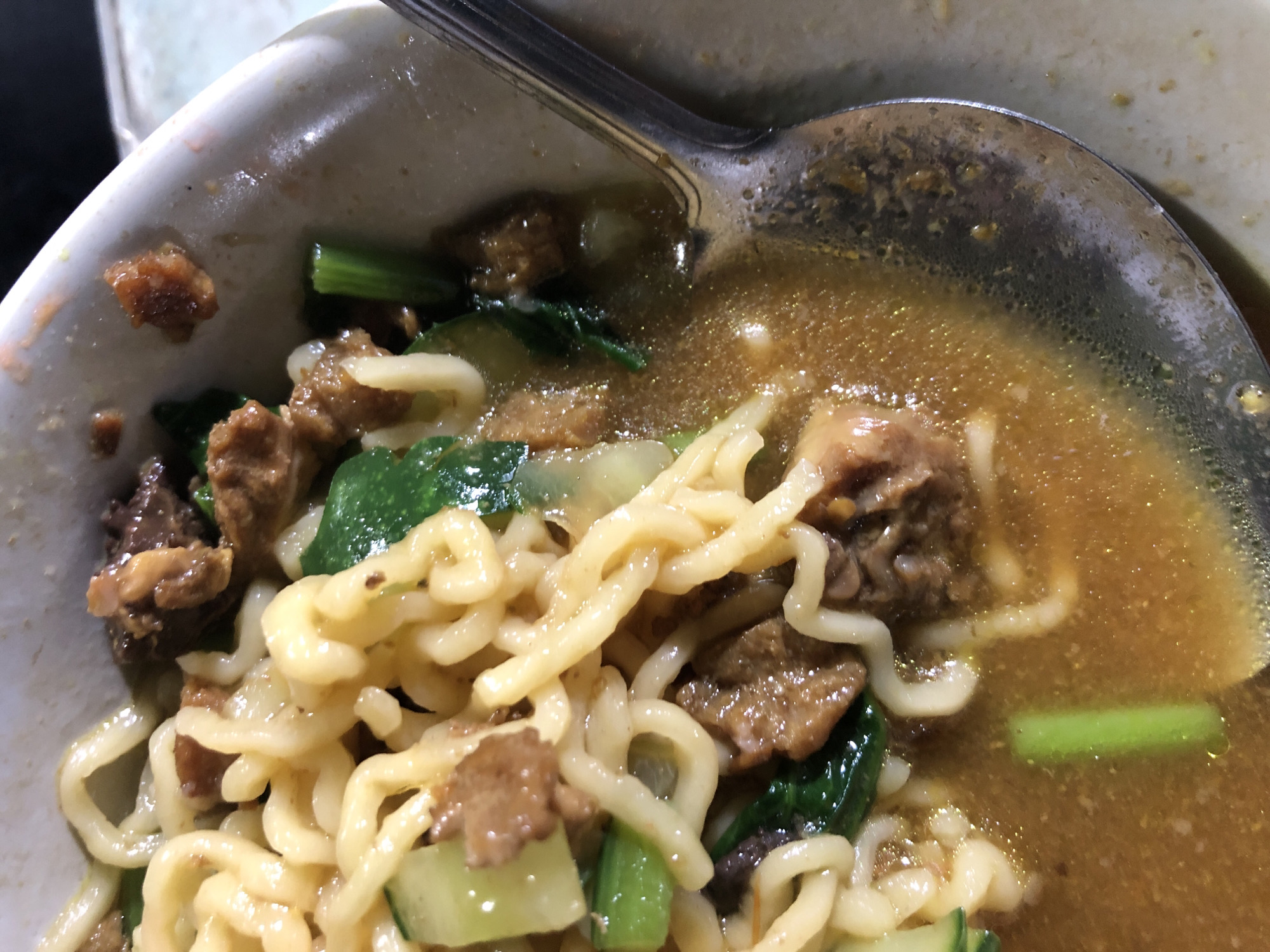 A close-up, overhead shot shows a bowl of noodles with broth and meat. The noodles are yellow and curly, and they are mixed with pieces of brown meat and green vegetables. A silver spoon is visible on the right side of the bowl, submerged in the broth. The bowl appears to be white and slightly chipped on the rim. The lighting is dim, and the background is out of focus, suggesting an indoor setting. The scene is likely in Sei Mencirim, Indonesia, given the context of the query.