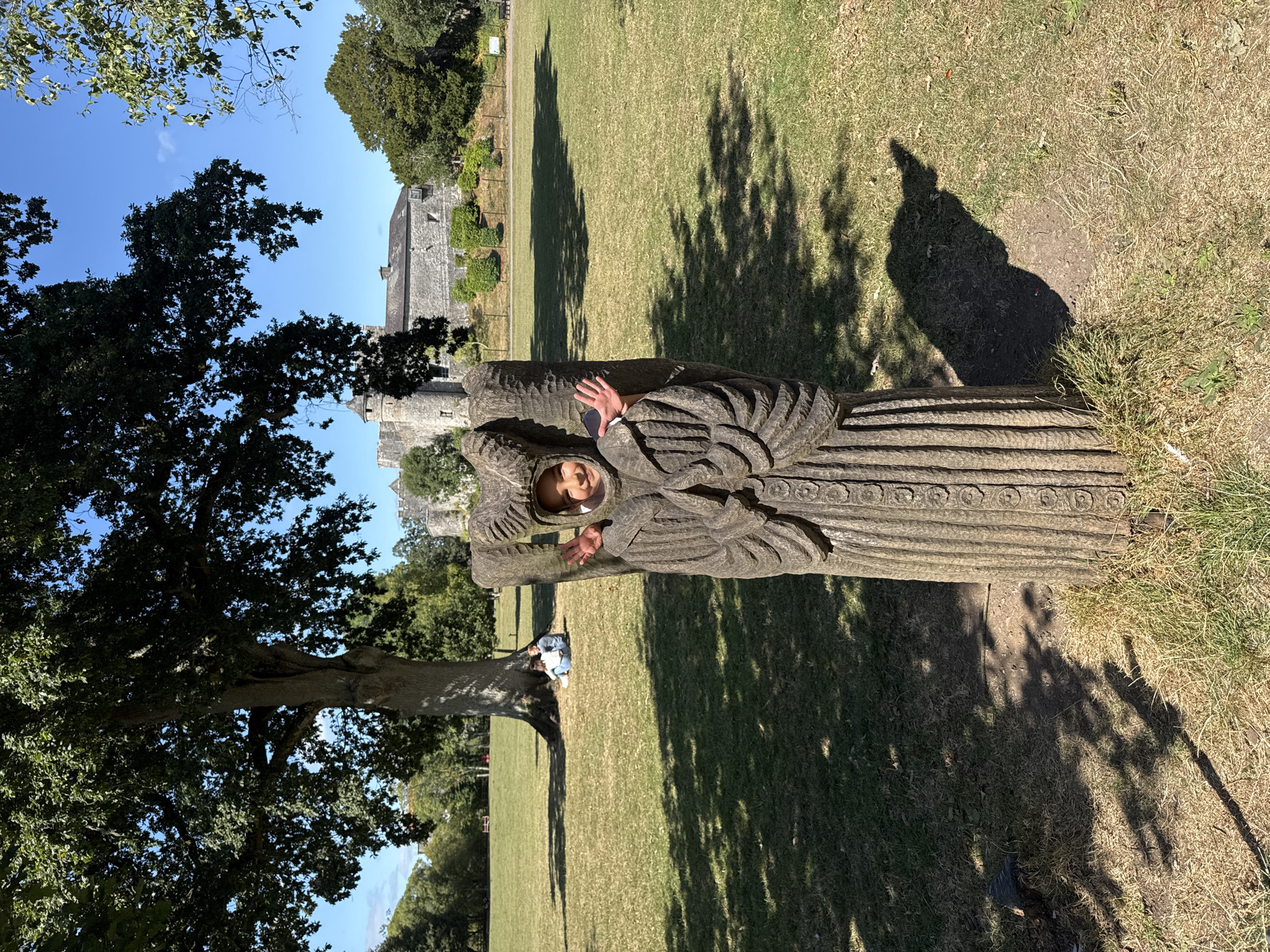 A young child with dark hair, wearing a light-colored top, peeks through a cutout in a large, weathered stone sculpture. The sculpture is carved to resemble a robed figure with intricate detailing and a headdress. The child smiles and waves with their right hand. The sculpture stands on a grassy field with dappled sunlight filtering through the leaves of a large tree behind it. In the background, a stone castle with a turret is visible, situated on a slight incline. Two other children sit on the grass near the base of the large tree. The scene appears to be outdoors in a park-like setting.