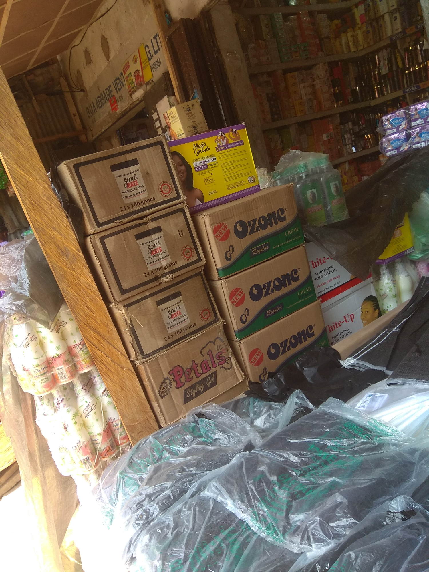 The image depicts a stall in the city of Jalingo, Nigeria, likely a market or small shop. The stall is filled with a variety of consumer goods, primarily cosmetics and personal care products. In the foreground, stacked cardboard boxes with branding such as "Soul Mate" and "Ozone" are visible, along with a prominent box labeled "Petals Styling Gel." Several plastic-wrapped bundles of what appear to be hair products or accessories are also present, along with large, dark-colored items enclosed in clear plastic sheeting, suggesting bulk goods or inventory.
In the background, shelves are laden with numerous bottles and packages of various brands, including NIVEA products. A partially visible sign at the top of the stall reads "BUA ABBARE VENT G. LTI". The environment appears somewhat cluttered and disorganized, typical of a bustling market setting. No people are clearly depicted in the image. The lighting suggests it is daytime.