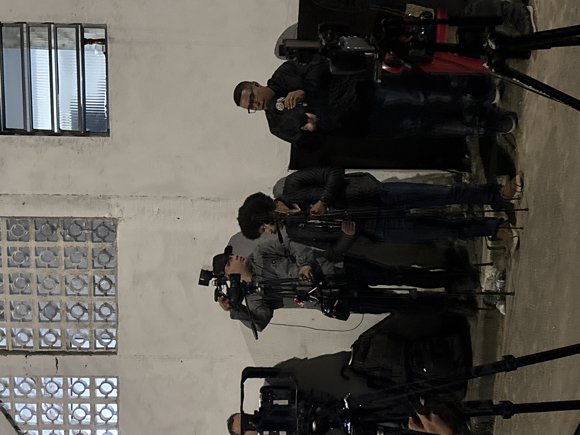 A group of people, primarily cameramen, are gathered outside a building with a window and a wall made of decorative blocks. The cameramen are operating video cameras mounted on tripods. One man in glasses appears to be giving an interview or statement while holding a microphone. This scene takes place in an urban area. It is not possible to definitively confirm the city of São Paulo, Brazil without more contextual information.