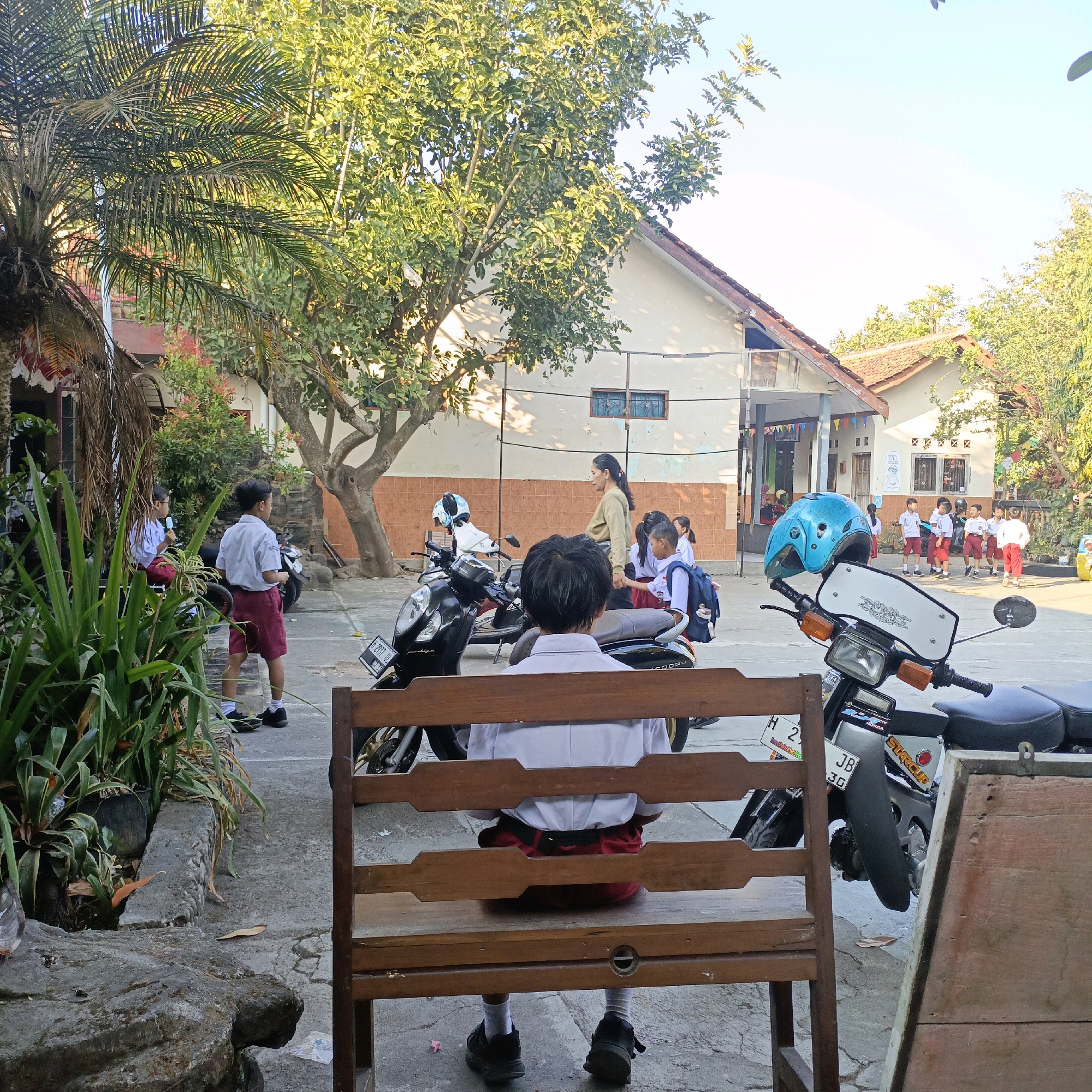 This is a photograph of a schoolyard in Salatiga, Indonesia, on a clear, sunny day. In the foreground, a young student wearing a white shirt and red shorts is sitting on a wooden bench, facing away from the camera. The student is seen from behind, with their back to the viewer. Beyond the bench, several motorcycles are parked. In the middle ground, a group of children in similar school uniforms are gathered, some with backpacks. An adult woman, possibly a teacher, is interacting with them. The background shows a building with a red tile roof, typical of Indonesian architecture, surrounded by trees and vegetation. The scene appears to be during school hours, possibly before or after classes, with children waiting or socializing. The environment is outdoors, with paved ground and a clear sky.