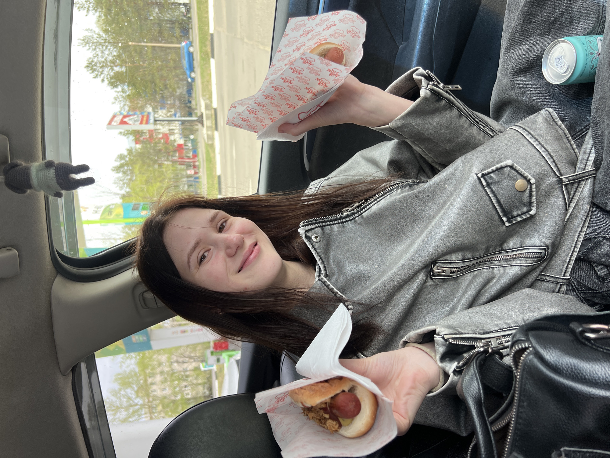 A young woman is seated in what appears to be the passenger seat of a car. She is holding two hot dogs wrapped in paper. The exterior view through the car window shows a street scene with buildings, signs, and trees. A small knitted toy is hanging near the window. A can of a beverage sits atop the woman's jacket.