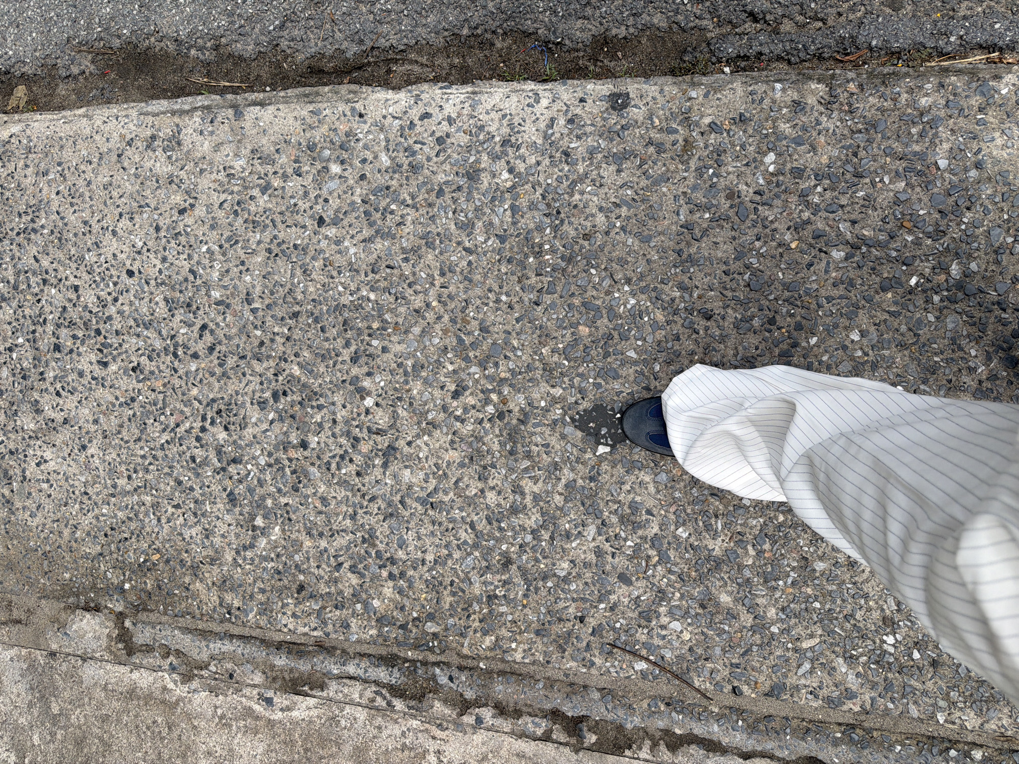 The image shows a close-up, overhead view of a person's lower leg and foot walking on a concrete sidewalk. The sidewalk is textured with small grey and black pebbles and has a raised edge. To the right of the sidewalk is a strip of darker, rougher asphalt. The person is wearing white pants with thin blue pinstripes and dark blue shoes. The scene appears to be outdoors, possibly in an urban environment, given the presence of a sidewalk and asphalt. The lighting suggests it is daytime. The location in Bangkok, Thailand, is not explicitly identifiable from the visual cues in the image.