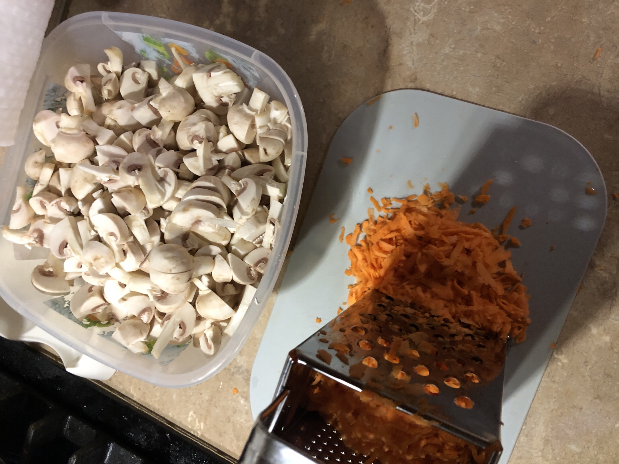 The image shows a kitchen scene with two main food items being prepared. In the upper right quadrant, a clear plastic container is filled with sliced white mushrooms. Below and to the left of the mushrooms, a metal grater is angled over a light gray cutting board. Shredded carrots are piled on the cutting board, having been grated from the grater. The countertop appears to be a speckled brown material. To the far left, a black stovetop is partially visible. The overall impression is of ingredients being prepped for cooking.