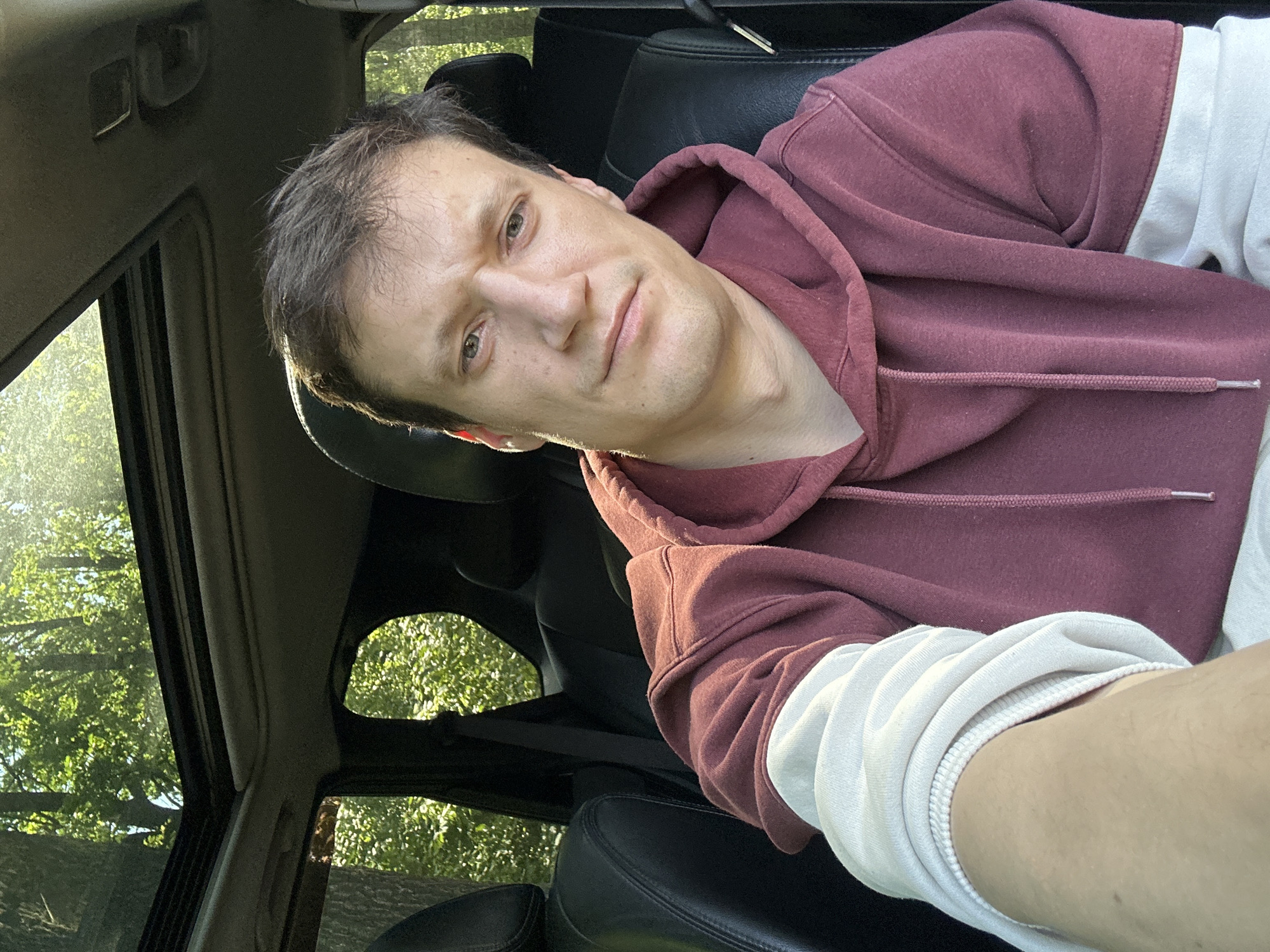 A man sits in the driver's seat of a car, looking at the camera. The car has a sunroof, through which sunlight streams in, illuminating the interior. The man is wearing a maroon hooded sweatshirt with white sleeves. The interior of the car appears to be black leather. Outside the car, through the windows and sunroof, lush green trees are visible, suggesting a wooded or natural environment. The scene does not contain any discernible elements that would indicate a specific location within the city of Makiv, Ukraine.