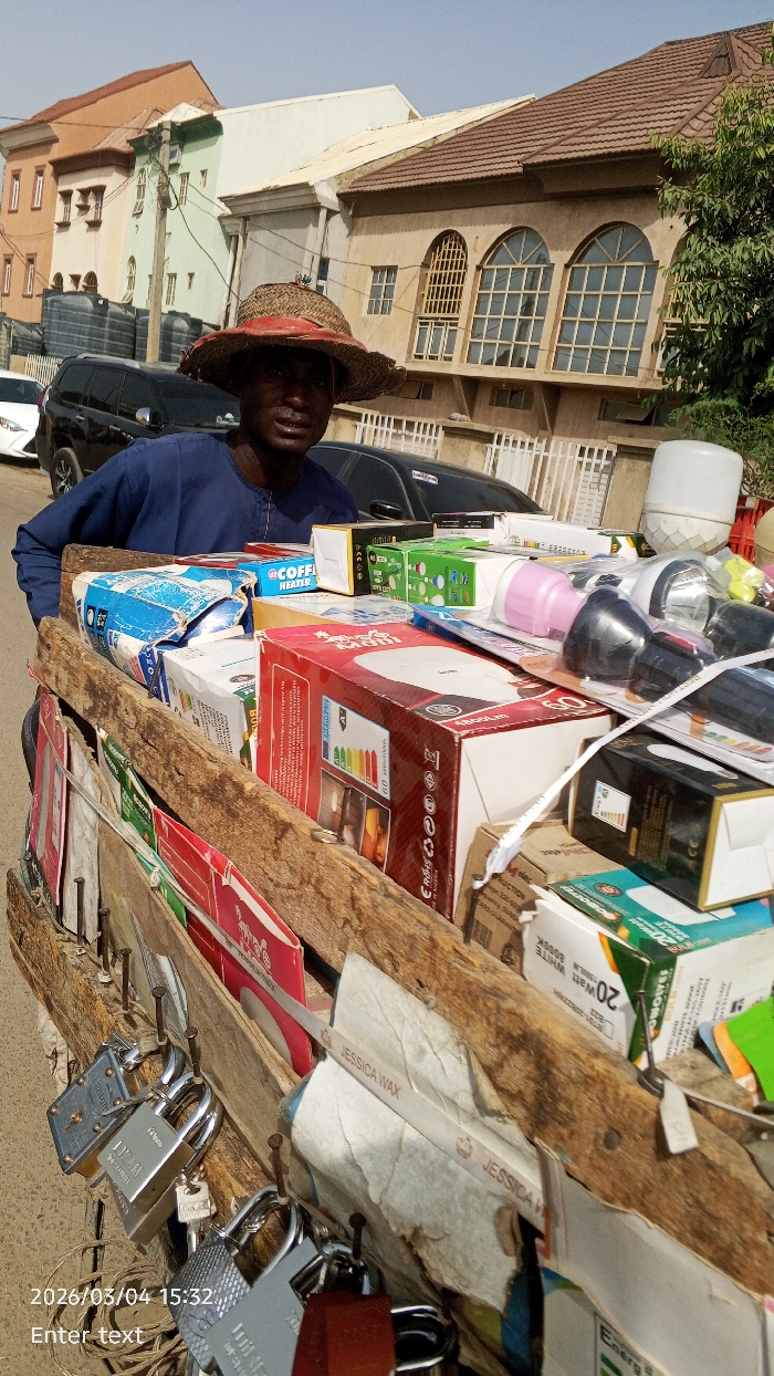 The image displays an outdoor street scene in Fagge A, Nigeria, timestamped 2026/03/04 at 15:32. A male individual, appearing to be an adult of African descent, is centrally positioned, looking directly at the camera. He wears a dark blue long-sleeved garment and a wide-brimmed straw hat with a red accent.
He stands behind a wooden pushcart, which functions as a mobile vendor's stall. The cart's top surface is densely packed with various boxed consumer goods, including items such as "COFFEE HEATER" packaging, numerous light bulbs or LEDs in their packaging, and multiple flashlights. The visible side of the cart is equipped with multiple metal fasteners from which numerous padlocks of different sizes and metallic finishes (e.g., silver, brass) are suspended, some with keys. Other packaged items like "JESSICA WAX" are also visible on the cart's side.
The background reveals a street with several parked vehicles, including a dark-colored SUV and a white sedan. Behind these vehicles, a row of multi-story buildings lines the street. From left to right, these include a light orange building, a light green building with a white upper section, a plain white building, and a prominent beige-colored building with a reddish-brown tiled roof. The beige building features three large arched windows with distinct grid patterns on its upper floor. A white fence runs in front of this building, and a green-leafed tree is partially visible on the far right. The sky above is clear and light blue, indicating bright daylight conditions. The ground in the immediate foreground appears to be an unpaved or dirt surface.