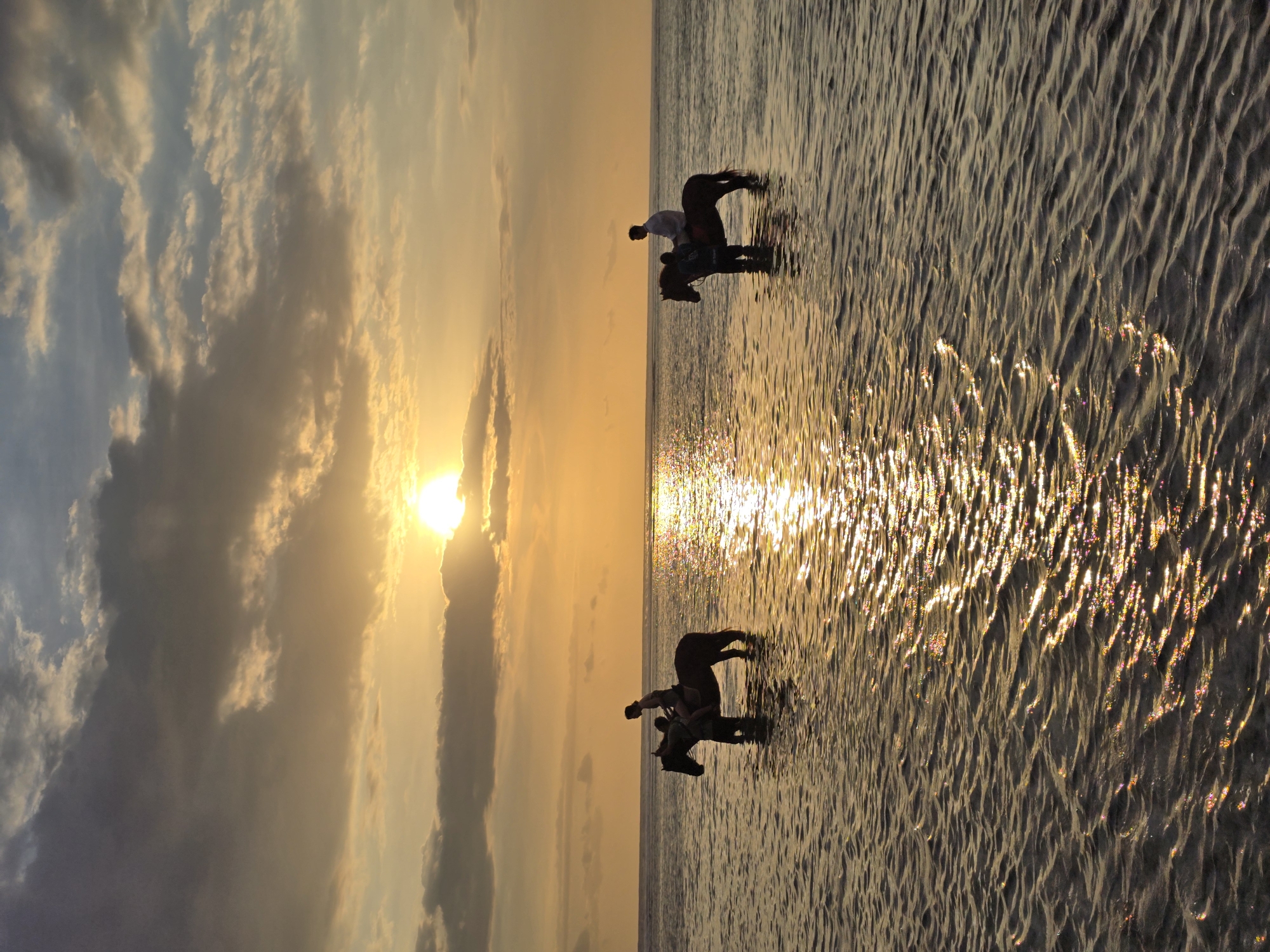 Here is a description of the image:
The image shows a tranquil coastal scene at sunset, likely in an Indonesian location. Two people are riding horses through shallow water. The water reflects the setting sun, creating a sparkling effect. The sky is filled with clouds, painted in warm hues of yellow and orange. The overall scene is peaceful and serene.