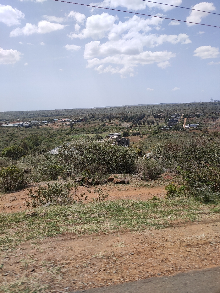 The image captures a wide, sunlit landscape view of what appears to be a rural or semi-rural area, consistent with Ewuaso Oonkidong'i ward, Kenya. The scene unfolds under a bright blue sky dotted with scattered white cumulus clouds.
In the foreground, a reddish-brown dirt path or ground stretches across the bottom, featuring sparse patches of green and dry grass and low-lying bushes. A dark, possibly asphalt, surface or car window ledge is visible at the very bottom edge of the frame.
The midground and background are dominated by an expansive, undulating terrain covered in dense natural vegetation, comprising a mix of green and dry bushes and trees, characteristic of a bushland or savannah environment. Scattered within this natural expanse are various buildings and small settlements. Some structures appear to be complete houses, while others, particularly in the midground, show exposed brickwork or concrete, suggesting ongoing construction or unfinished development. These buildings are spread out, indicating a low-density population.
Overhead, power lines are visible, stretching horizontally across the sky from left to right. In the far distance on the right, several tall electricity pylons or transmission towers stand against the horizon, indicating established infrastructure.
The time of day is clearly daylight, likely late morning or early afternoon, given the bright illumination and visible cloud patterns. No human activity is discernible, and no text is visible within the image. The overall impression is a vast, natural landscape with evidence of gradual human settlement and development.
