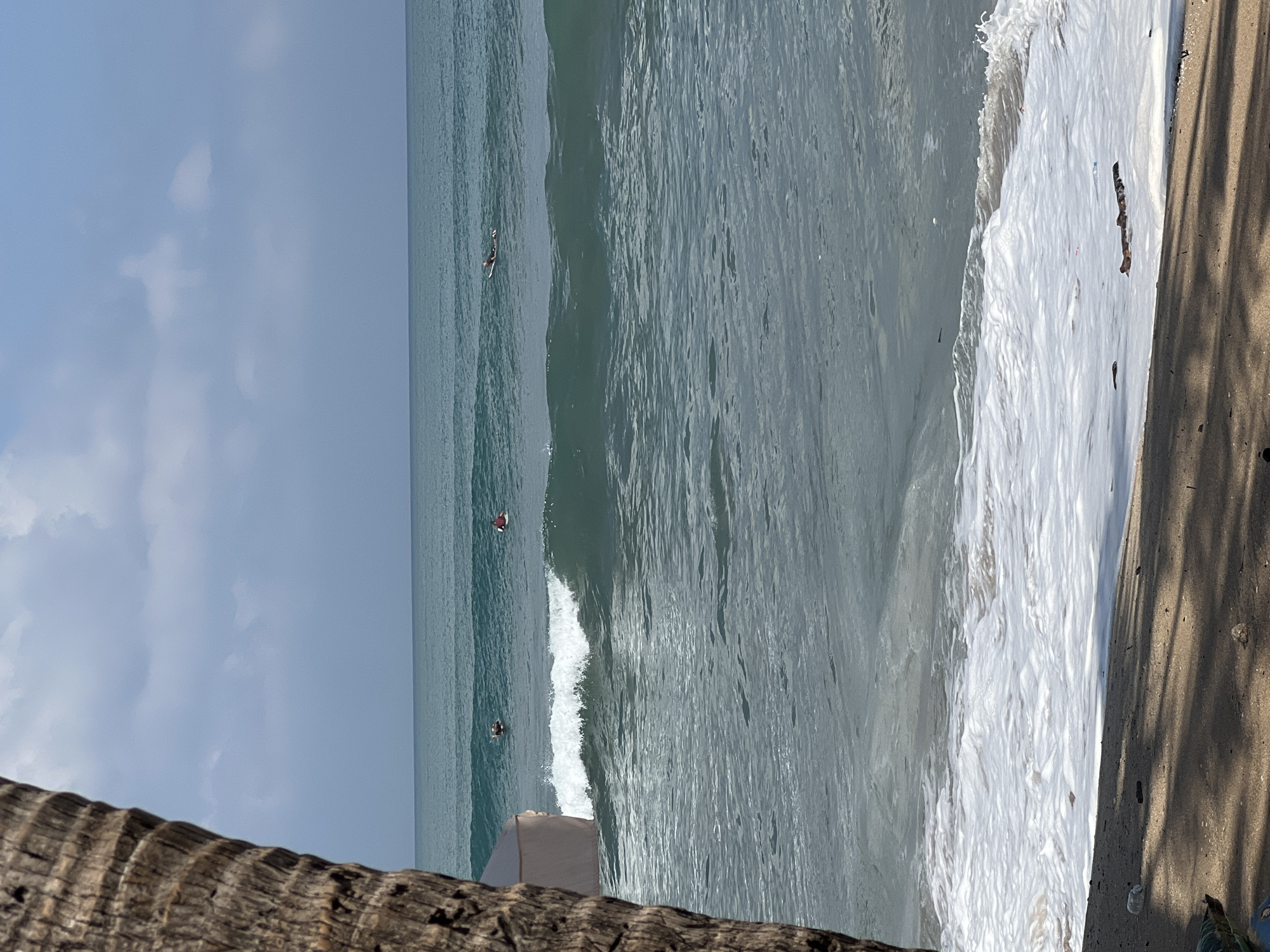 The image depicts a view of the ocean at Kuta, Indonesia. A portion of a palm tree trunk is visible in the foreground on the left side. The sea is calm with small waves breaking near the shore, creating white foam. Several surfers are scattered across the water, some riding waves and others paddling. The sky is partly cloudy with a pale blue hue. The beach in the lower portion of the image is sandy, with some debris and shadows cast by overhead foliage.