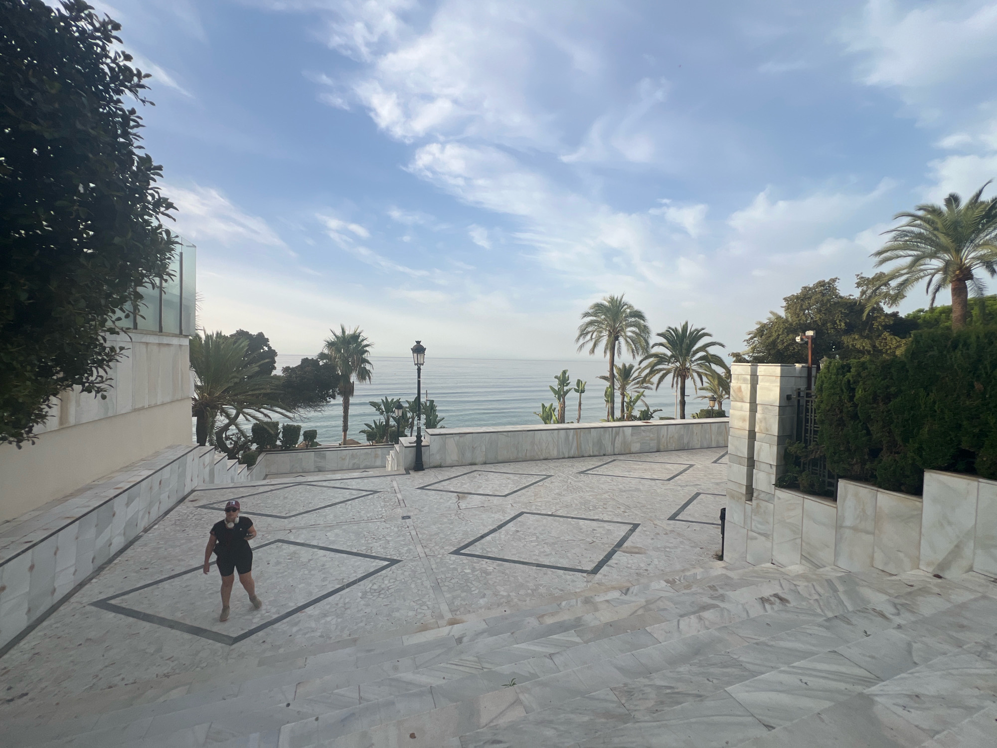 The image depicts a person walking on a patterned marble staircase that leads to a paved plaza overlooking the Mediterranean Sea in Marbella, Spain. Palm trees and lush greenery are visible in the background, with the calm sea stretching to the horizon under a partly cloudy sky. The architecture features white marble walls and accents, creating a clean and luxurious aesthetic.