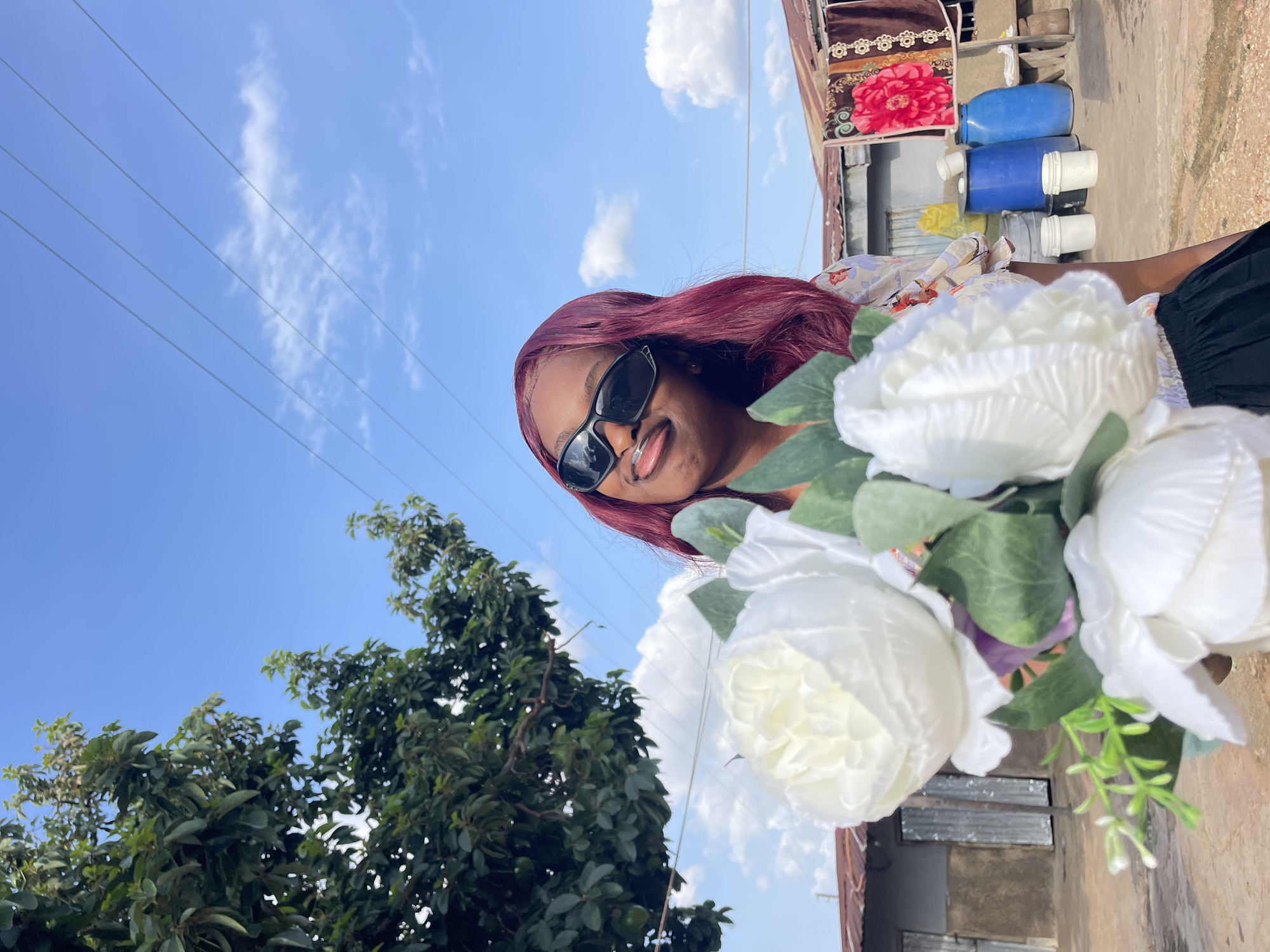 A woman with dark skin and long, reddish-brown hair is shown in a close-up, rotated view, holding a bouquet of white flowers. She is wearing dark sunglasses and has a playful expression, sticking her tongue out slightly. The background features a bright blue sky with wispy clouds, suggesting it is daytime and fair weather. Power lines are visible crisscrossing the sky. To the right of the woman, there are some objects stacked, including blue plastic containers and what appears to be a rolled-up blanket with a floral pattern. There is also some corrugated metal and brickwork visible, hinting at an outdoor setting, possibly a yard or an area with some structures. A large tree with lush green leaves is visible on the left side of the image. The overall mood is casual and cheerful. No visible text is present. The location context of Jos, Nigeria, is provided.