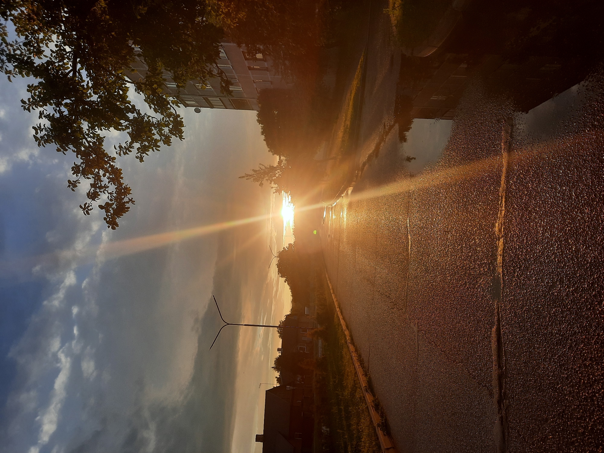 The image depicts a street scene in Paretcha, Belarus, during a sunset. The sun is low on the horizon, casting a strong, golden light and creating visible sun rays. The asphalt road is wet, with puddles reflecting the surrounding environment. On the left side of the road, there are several houses and trees. On the right side, a multi-story building is visible, partially obscured by trees. The sky is a mix of clouds and blue sky, with dramatic lighting from the setting sun. There are no people visible in the image. The overall impression is one of a quiet, residential street at the end of the day.