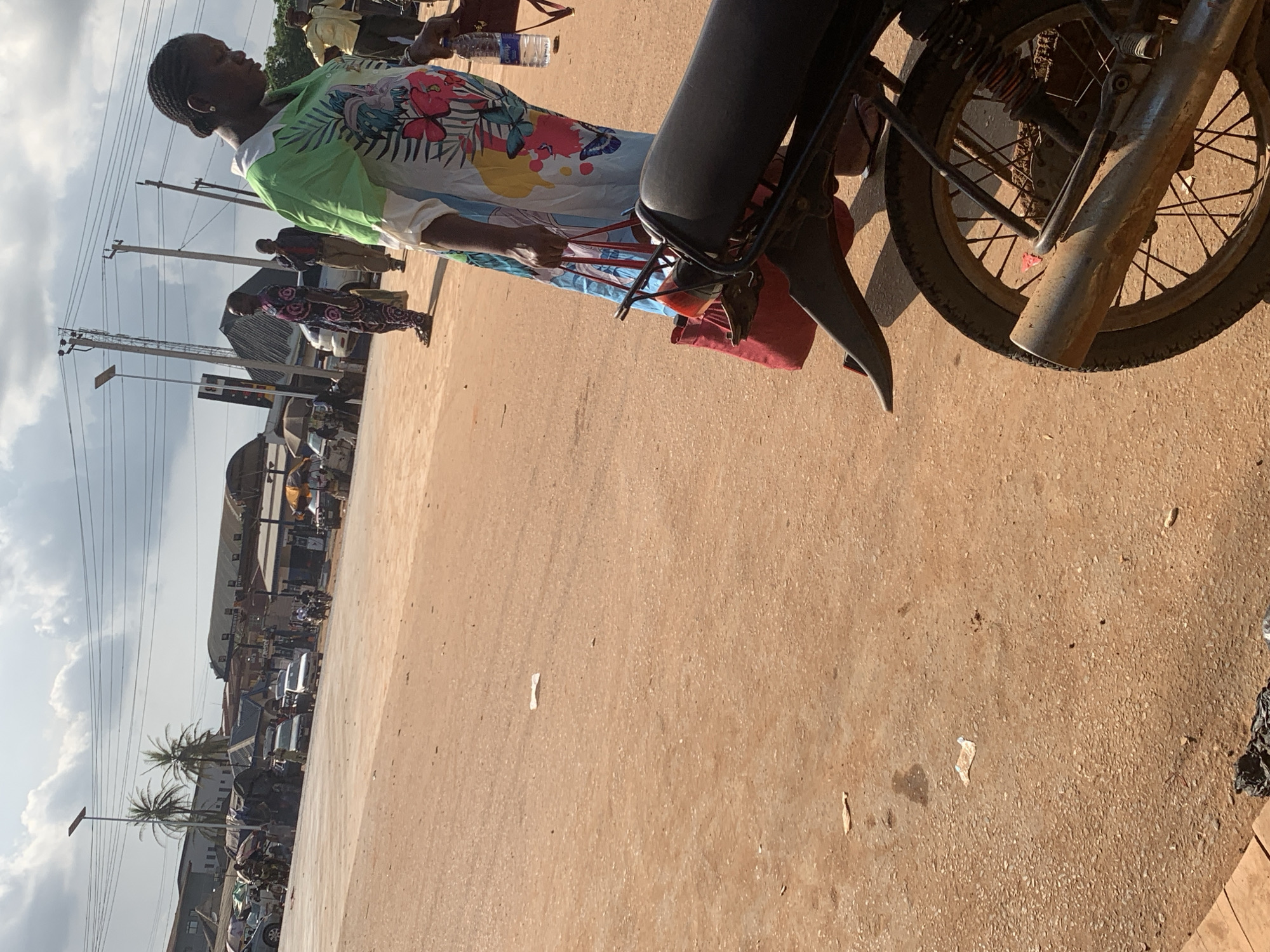 A person is standing near a motorcycle in a street scene in Shagamu, Nigeria. The person is wearing a colorful, patterned dress and has their hair in braids. They are holding onto the back of the motorcycle, which has a dark seat and a spoked wheel. In the background, there are other people, buildings, and vehicles, including cars and more motorcycles. Power lines and utility poles are visible against a cloudy sky, suggesting daytime. The ground appears to be a dusty, reddish-brown dirt or gravel road. The overall impression is of a busy street or market area.