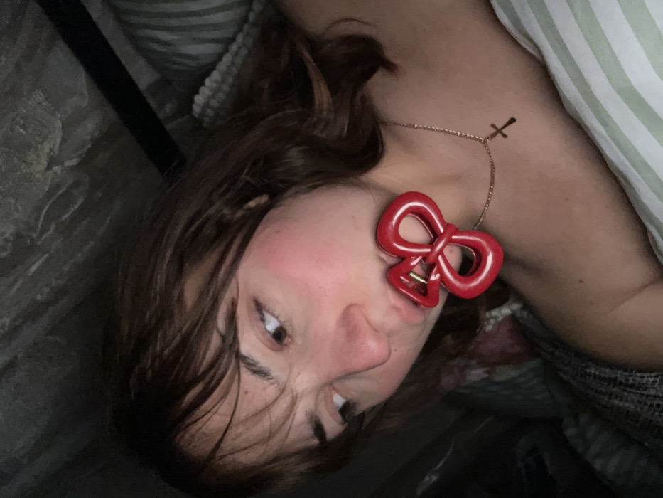 A woman lies on her side in bed, facing the camera. She has brown hair and her face is close to the lens. A red bow-shaped hair clip is attached to her eyebrow. She is also wearing a delicate gold necklace with a small cross pendant. The bedding includes a striped pillow and a floral-patterned blanket. The overall lighting is subdued, and the image is slightly rotated.