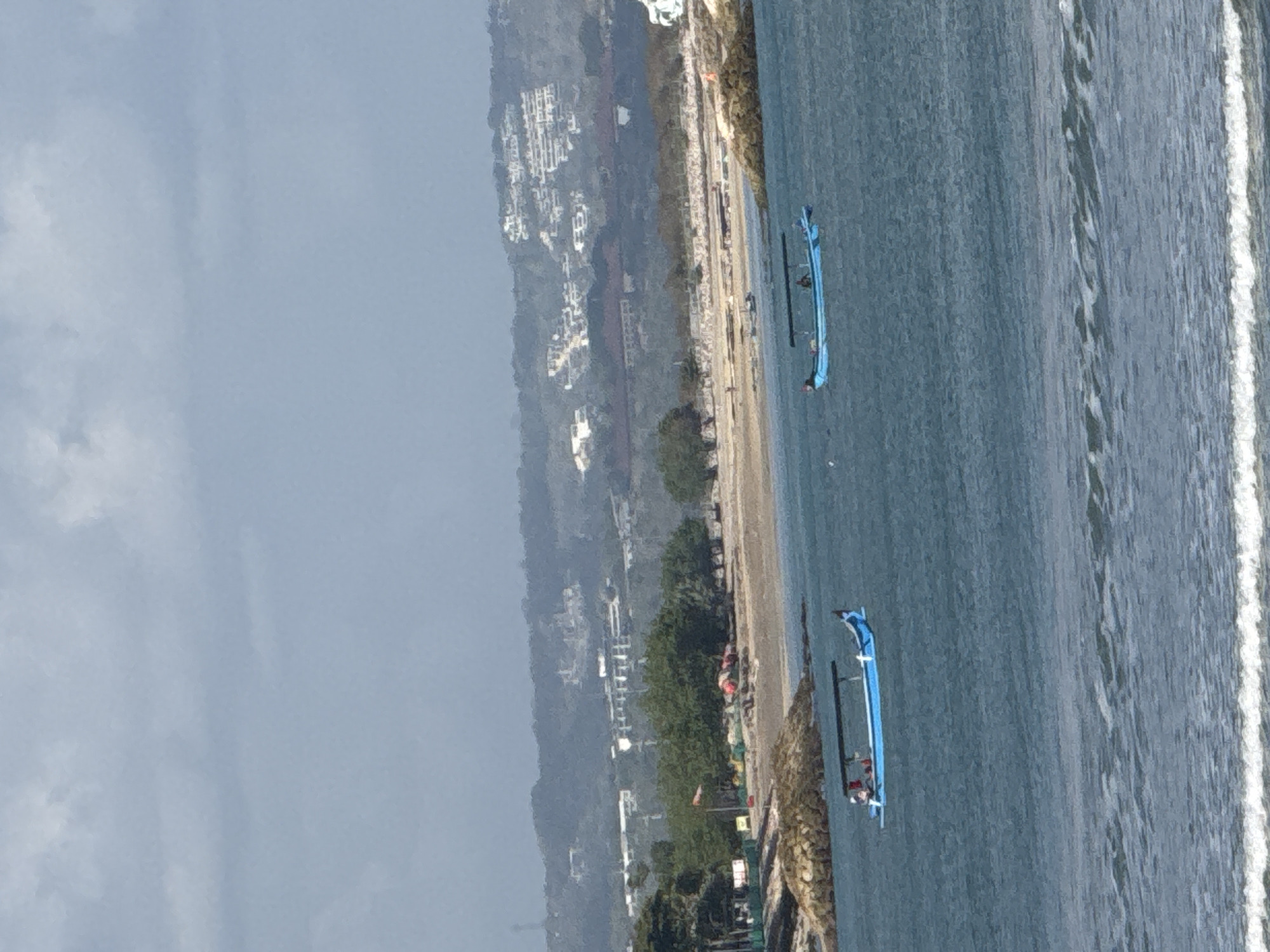 The image depicts a coastal scene with two blue boats in the foreground, navigating through calm, blue waters. One boat, closer to the viewer and on the left side of the frame, carries at least two individuals. The second boat, further out and on the right, appears to have one person aboard. In the midground, a sandy beach is visible, dotted with trees and some structures, including what appears to be a windsock indicating a public area. Rocky outcrops break the shoreline. Beyond the beach, a hillside is covered with a dense arrangement of white buildings, suggesting a populated area with resorts or residential complexes. The sky above is overcast with muted grey clouds. The scene is consistent with the coastal environment of Kuta, Indonesia, a popular tourist destination known for its beaches and seaside development.