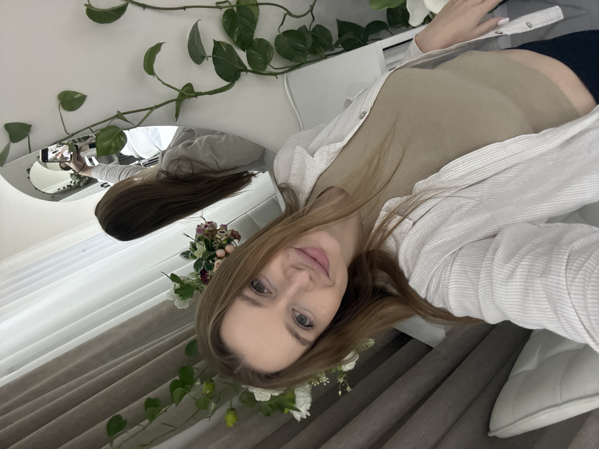 An indoor scene in Budva, Montenegro, depicts a female capturing a selfie in an arched mirror. The subject, a woman with long light brown hair and light-colored eyes, is wearing a light tan ribbed tank top beneath an unbuttoned light-striped long-sleeve shirt and dark bottoms. She holds a black smartphone in her raised right hand, directing it towards the mirror, with her own image visible on the phone's screen. Her left hand, featuring white polished nails, is visible near her hip. The immediate environment includes a dark, textured curtain on the left, layered with sheer white curtains. Green leafy vines are arranged on the white wall to the right and near the curtains, complemented by white and pink/burgundy floral arrangements. A white cushion is visible in the bottom left foreground, and white furniture is reflected in the mirror behind the subject.