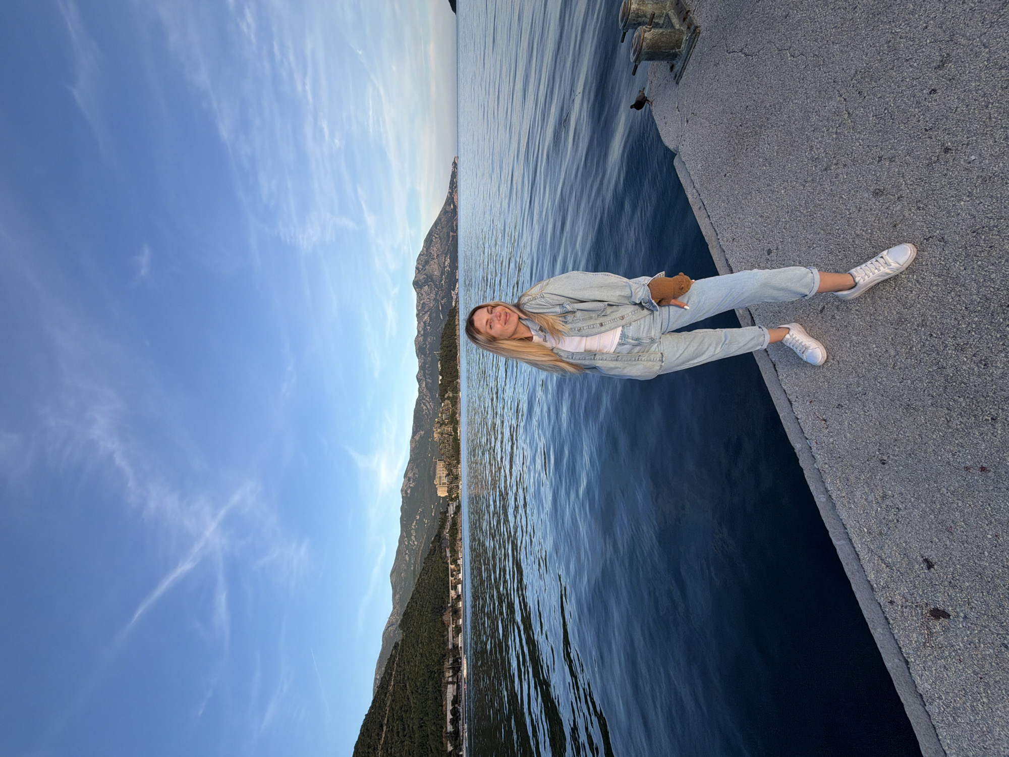 A young woman with blonde hair sits on a concrete pier alongside the Adriatic Sea. She wears a denim jacket and jeans with white sneakers and holds a small, brown teddy bear in her lap. The sea is calm, and the mountainous coastline of Montenegro is visible in the background. The sky is blue with wispy clouds. The image was likely taken in or near Budva, Montenegro, given the coastal and mountainous landscape.