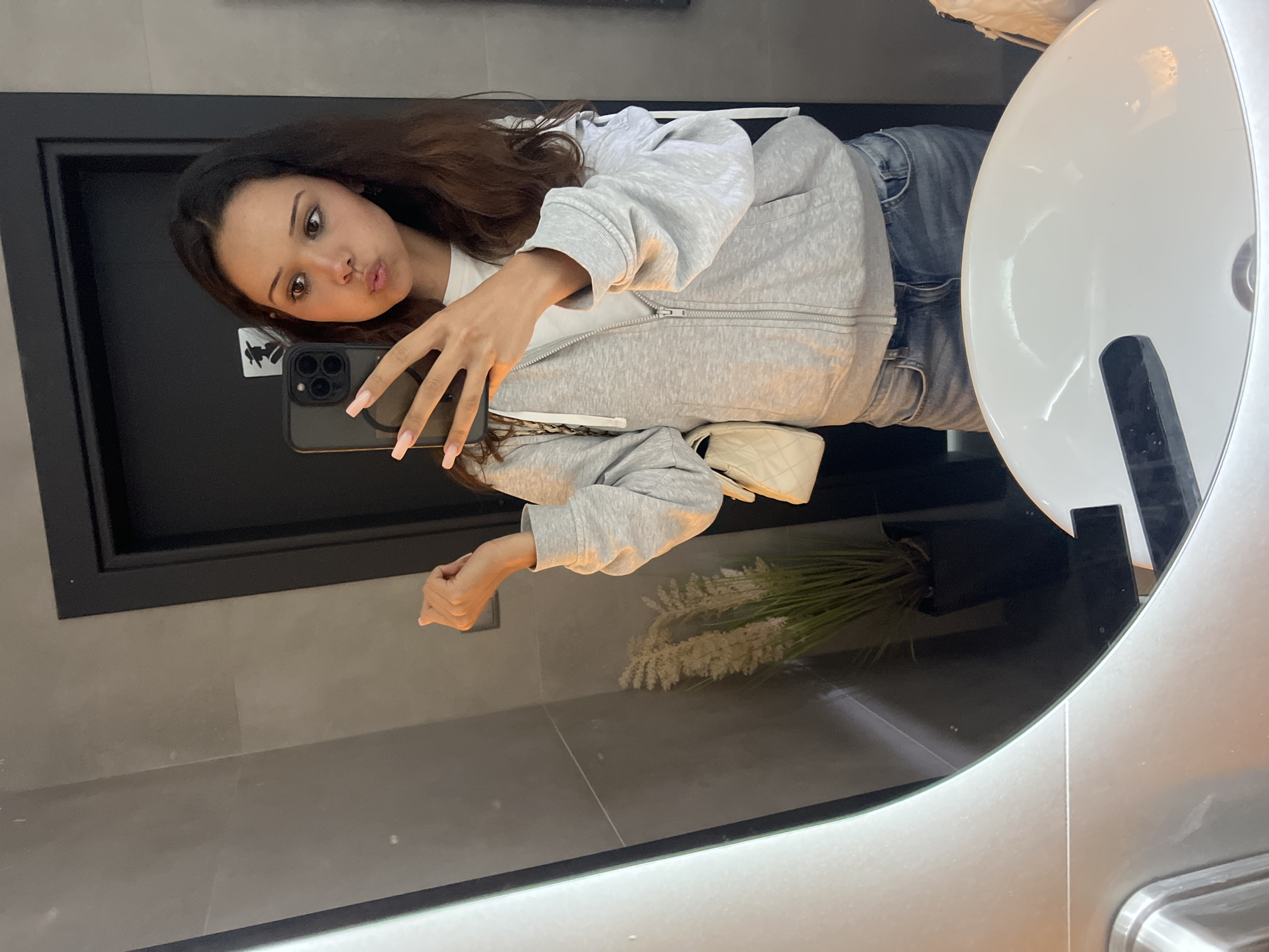 In Zlatograd, Bulgaria, a young woman with brown hair and light skin is taking a mirror selfie in what appears to be a public restroom. She is wearing a grey zip-up hoodie over a white t-shirt and blue jeans. Her nails are painted a light pink. She is holding a smartphone in her right hand, with her left hand resting on the side of the mirror. In the background, a potted plant with dried grass-like stalks is visible, along with a dark door and grey tiled walls. A white purse with a quilted texture is hanging from her left shoulder. The sink in the foreground is white and modern in design.