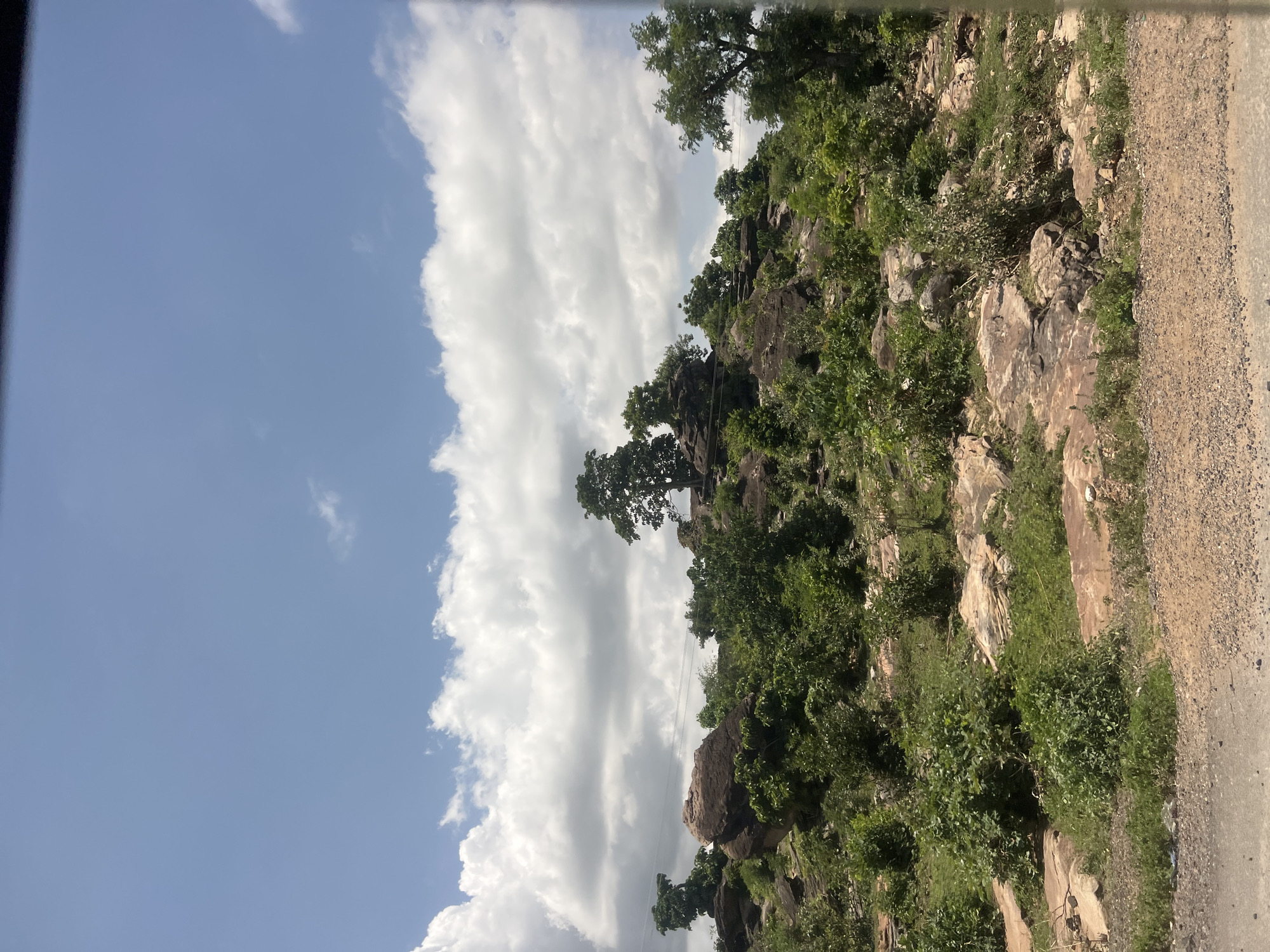 This image displays a natural, rocky hillside landscape under a partly cloudy blue sky in Lafia, Nigeria. The upper portion of the frame features a clear blue sky interspersed with large, white cumulus clouds. The terrain consists of an upward-sloping expanse of numerous dark brown and reddish-brown boulders and exposed rock formations. Patches of green shrubbery and trees are densely scattered across the rocky ground, with several larger trees prominent on the higher elevations. Multiple thin, dark power or communication lines stretch horizontally across the mid-ground, passing above the vegetation and rocks. The extreme bottom of the image reveals a narrow strip of a dirt or gravel road, suggesting a roadside viewpoint. No people or animals are visible in the scene.