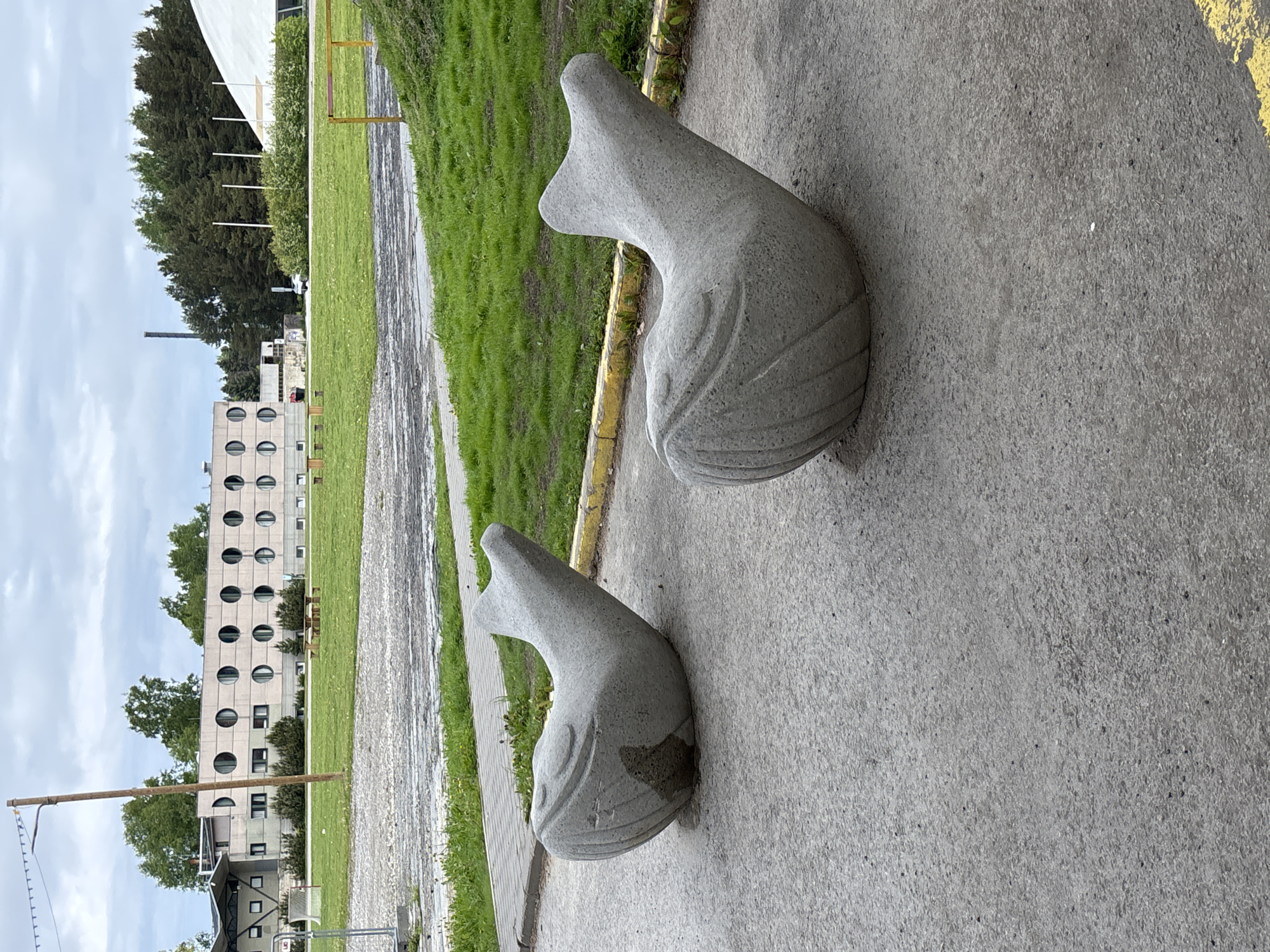 The image shows two stone whale-shaped sculptures sitting on a paved area. They are gray in color with carved details. In the background is a grassy area and a building with round windows, indicative of architecture in Haabneeme alevik, Estonia. The sky is overcast. There are no people visible.