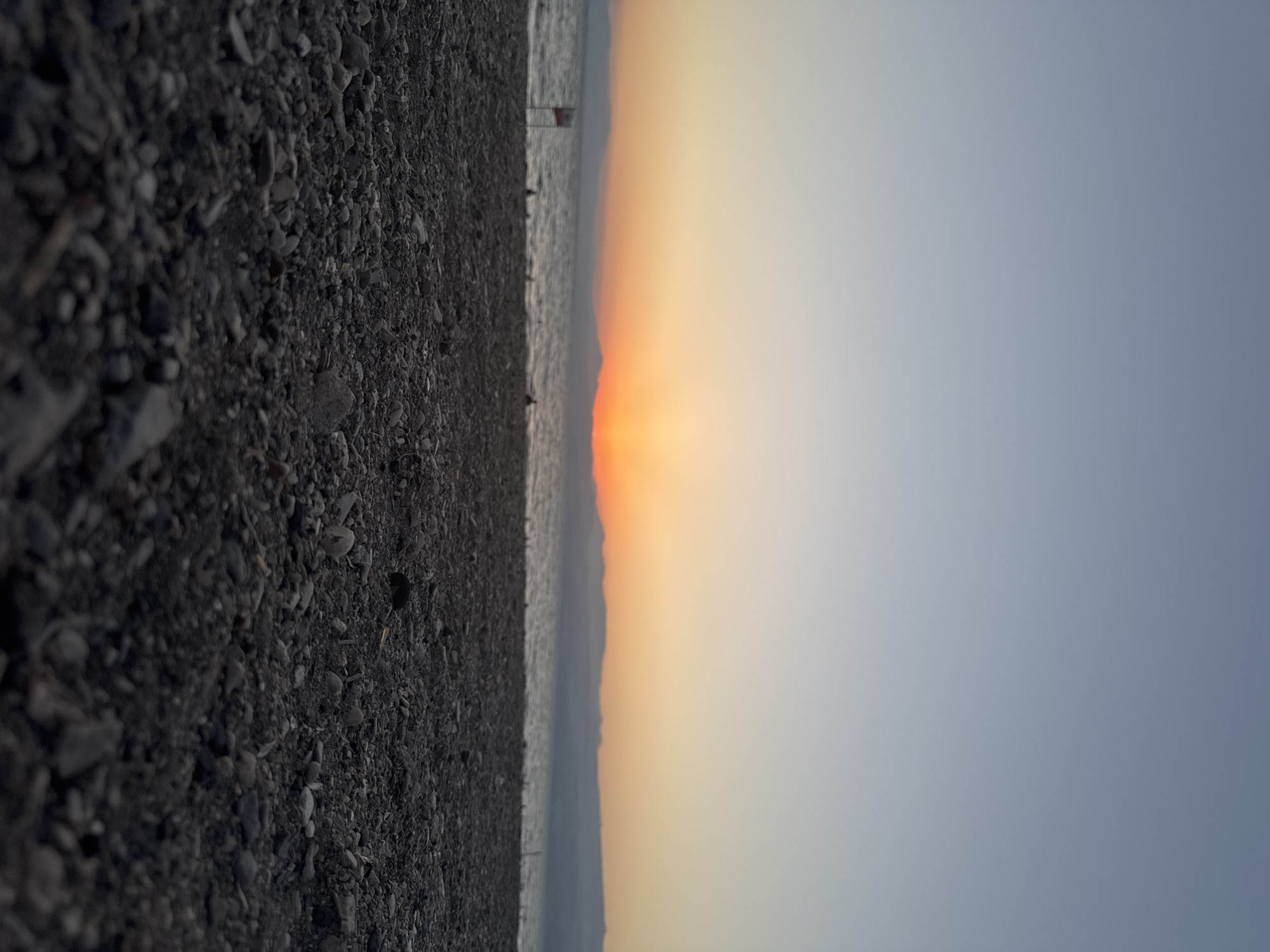 The image depicts a low-angle view of a rocky beach at sunset in the Golan Regional Council, Israel. The foreground is dominated by dark, gray pebbles and stones. In the mid-ground, a body of water, likely the Sea of Galilee, is visible with a distant, blurred shoreline featuring hazy mountains. The sky transitions from a pale blue at the top to warm orange and yellow hues near the horizon, indicating sunset. A small, blurred red and white sign is present on the left side of the beach, near the water's edge. Several indistinct, blurred figures are scattered along the shoreline. The overall impression is one of a serene, natural landscape at dusk.