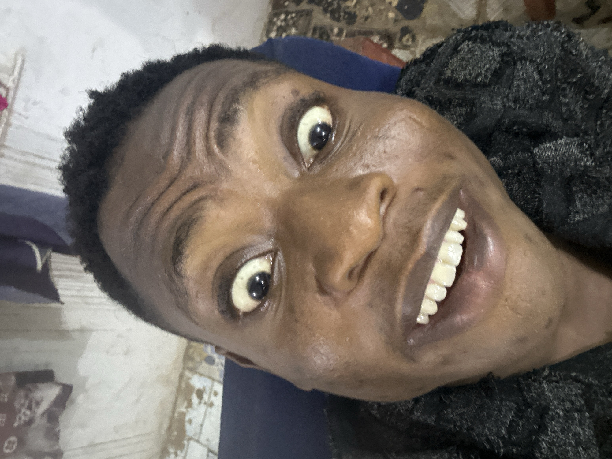 A close-up, angled portrait captures a dark-skinned individual with short, dark hair and a wide, toothy grin. Their eyes are wide and expressive. The person appears to be wearing a dark-colored, textured top. The background is somewhat blurry and indistinct, showing a floor.