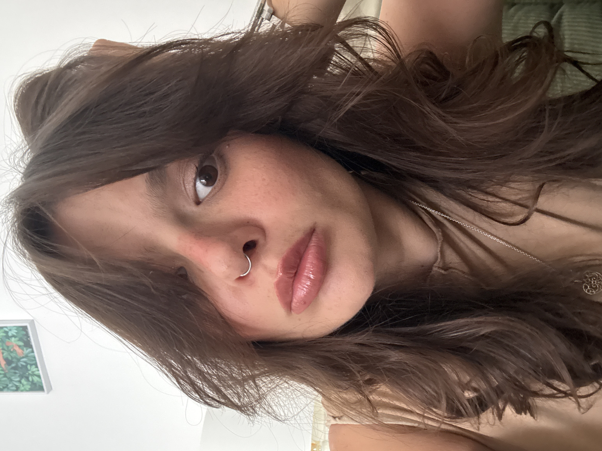 A close-up, high-angle portrait captures a young woman, likely indoors in Tel-Aviv, Israel, during the daytime. Her head is tilted, with her face occupying the central and lower-right portions of the frame. She has long, wavy, medium-brown hair that flows around her face and over her shoulders, partially obscuring her left eye. Her right eye is clearly visible, light brown or hazel, and she gazes directly at the viewer. A small, silver hoop nose ring adorns her left nostril. Her lips are full and glossy, tinted with a nude or light pink shade. A faint, natural blush is visible on her cheeks. Around her neck, she wears a thin silver chain necklace with a small, decorative silver pendant, possibly featuring a filigree design. The background is softly lit, showing a plain white wall on the left with a partially visible framed artwork depicting green foliage. On the upper right, a green-toned, subtly striped fabric object, possibly a cushion or headboard, is visible. The woman's expression is neutral and direct. There is no visible text in the image.