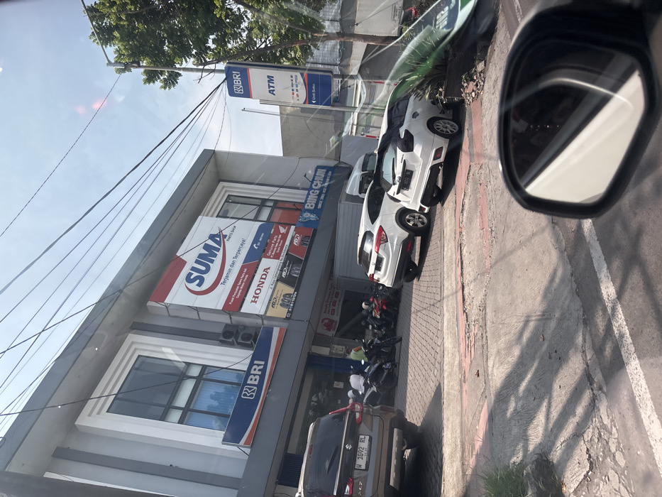 The image shows a street scene in Sidoarjo, Indonesia (latitude: -7.442, longitude: 112.687). A gray brick building with a large sign advertising "SUMA Parts Shop" is visible. The sign is red with white text, and there are two smaller signs for HONDA and BRI underneath it. There is a white Honda HR-V car parked in front of the building, and another white car, possibly a Toyota Yaris, is parked behind it. There are several motorcycles parked in front of the building, including some with people sitting on them. The street is made of gray concrete and has a white line painted down the center. There is a large tree on the right side of the street, and a sign for BRI ATM is visible in the image. There are also a few people walking on the sidewalk. The sky is a light blue with some clouds. The scene is shot from the perspective of a car traveling down the road. The car's mirror is visible in the lower right corner of the image.