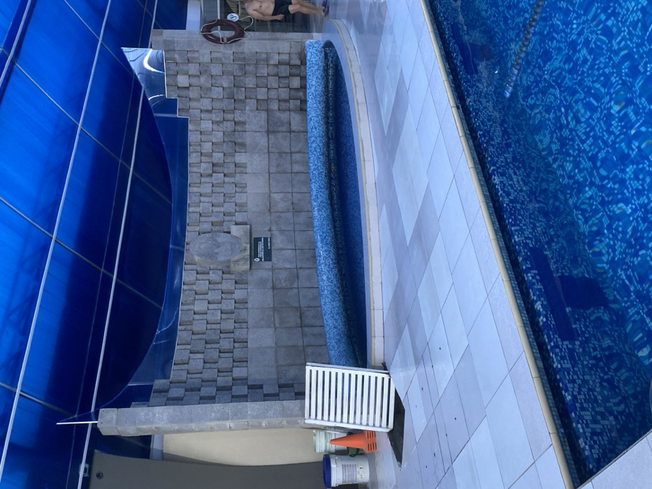 A top-down shot shows a swimming pool and its surroundings. The pool's edge is tiled in white, leading to a blue-tiled pool surface. A small, curved, blue-tiled section parallels the pool. Next to the pool is a tiled walkway leading to a blue structure. The walkway features various tile patterns. A lifeguard float, some construction materials, and a grate are near the walkway. A person is relaxing on a lounge chair in the distance.