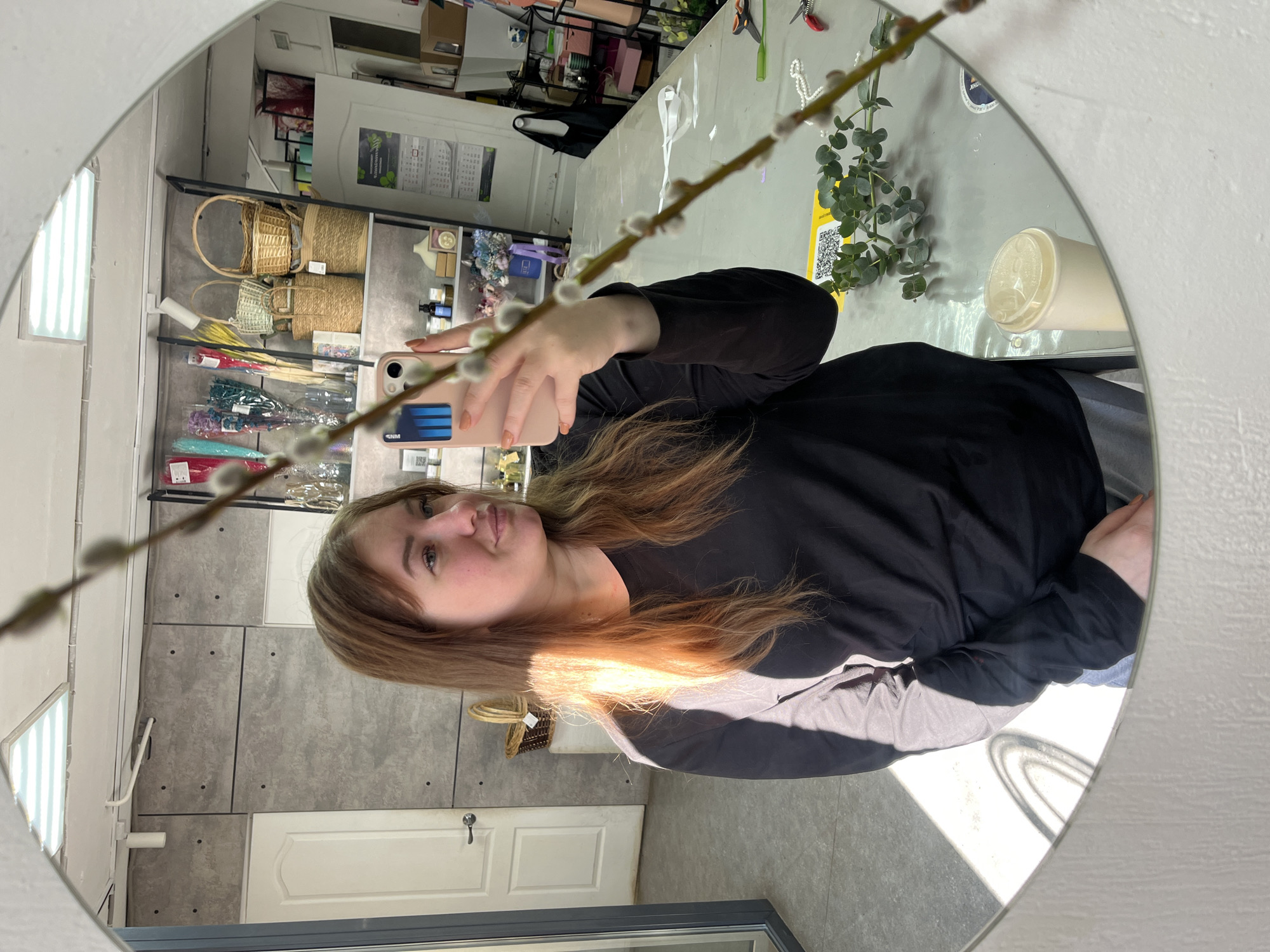 A selfie taken in a mirror shows a young woman holding a smartphone, reflected in a mirror. She is wearing a black long-sleeved shirt. In the background, shelves display baskets and other items, suggesting a retail environment. There is also a white door and grey-toned wall. The reflection shows a table with plants and other objects. While a general description can be provided, it is not possible to confirm that the location is in Chaikovsky, Russia based on only an image.