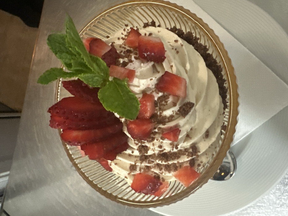 The image displays a dessert presented in a clear, ribbed glass bowl with a gold-colored rim, located in the city of Razlog, Bulgaria. The dessert is composed of a dark crumb base layered with a light-colored, swirled cream or mousse. Garnish includes several vertically sliced red strawberries positioned at the rear of the bowl, smaller diced red strawberry pieces scattered across the cream, dark brown sprinkles, and a sprig of fresh green mint leaves. The bowl is situated on a white, round plate, which rests partly on a folded white napkin visible at the lower right. A silver spoon handle is partially visible on the white plate at the lower left. The background features a blurred, reflective metallic surface, consistent with a stainless steel countertop found in a food preparation or service area. No individuals are present in the frame.