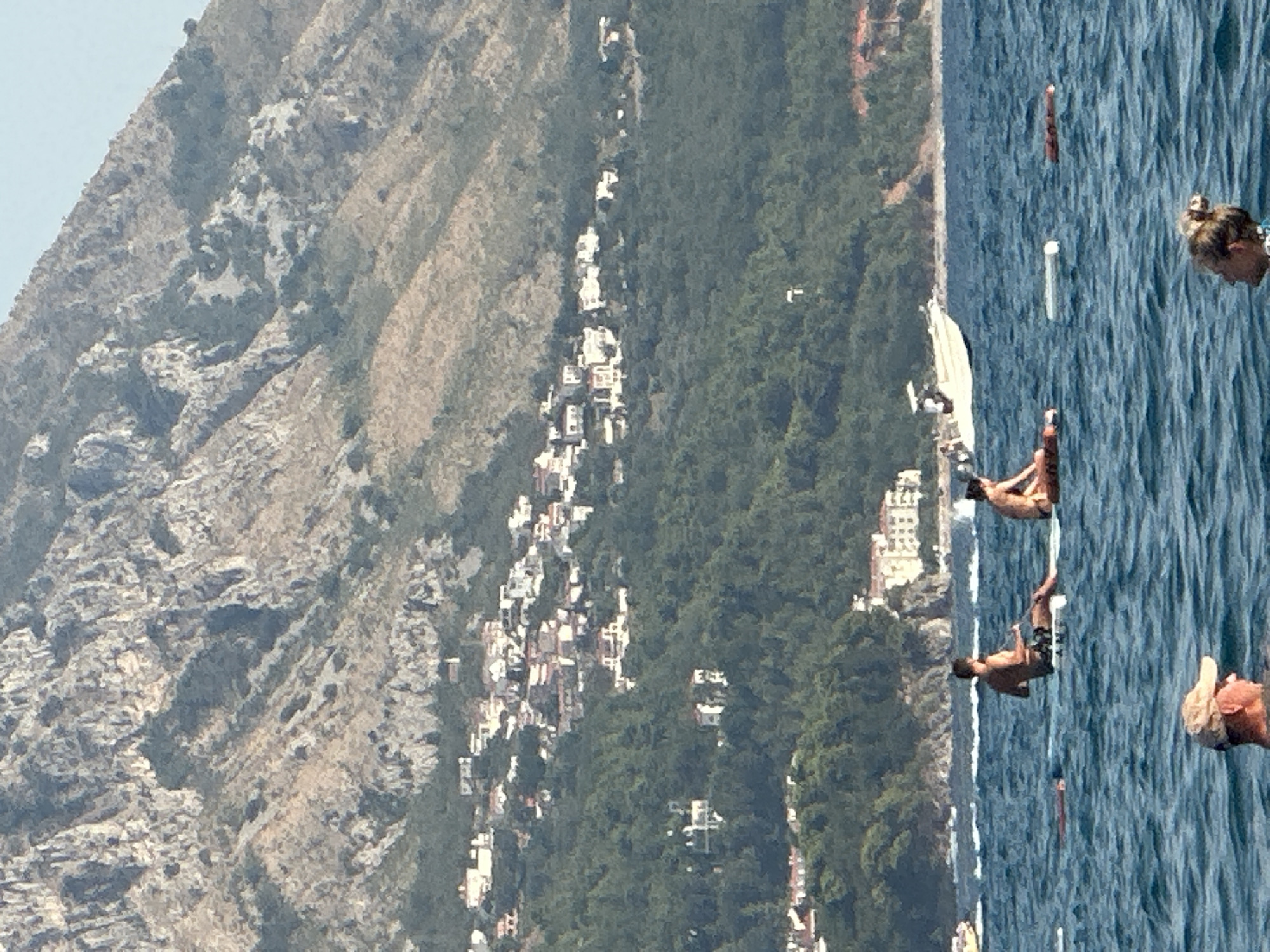 In the city of Boreti, Montenegro, this image captures a serene coastal scene with a mountainous backdrop. The water is a deep blue, with gentle waves rippling across the surface. In the foreground, a man wearing a cap is partially submerged in the water, facing to the right. Further out in the water, a young woman with her hair in a bun is also partially visible. Two individuals are seen on separate paddleboards. The man on the left paddleboard is seated and facing away from the viewer. The woman on the right paddleboard is seated and facing to the left. In the middle ground, a white speedboat with several occupants is visible. The shoreline features lush green vegetation and scattered buildings with red-tiled roofs, characteristic of Mediterranean architecture. Above the vegetation, a rocky, arid mountain range dominates the upper portion of the image.