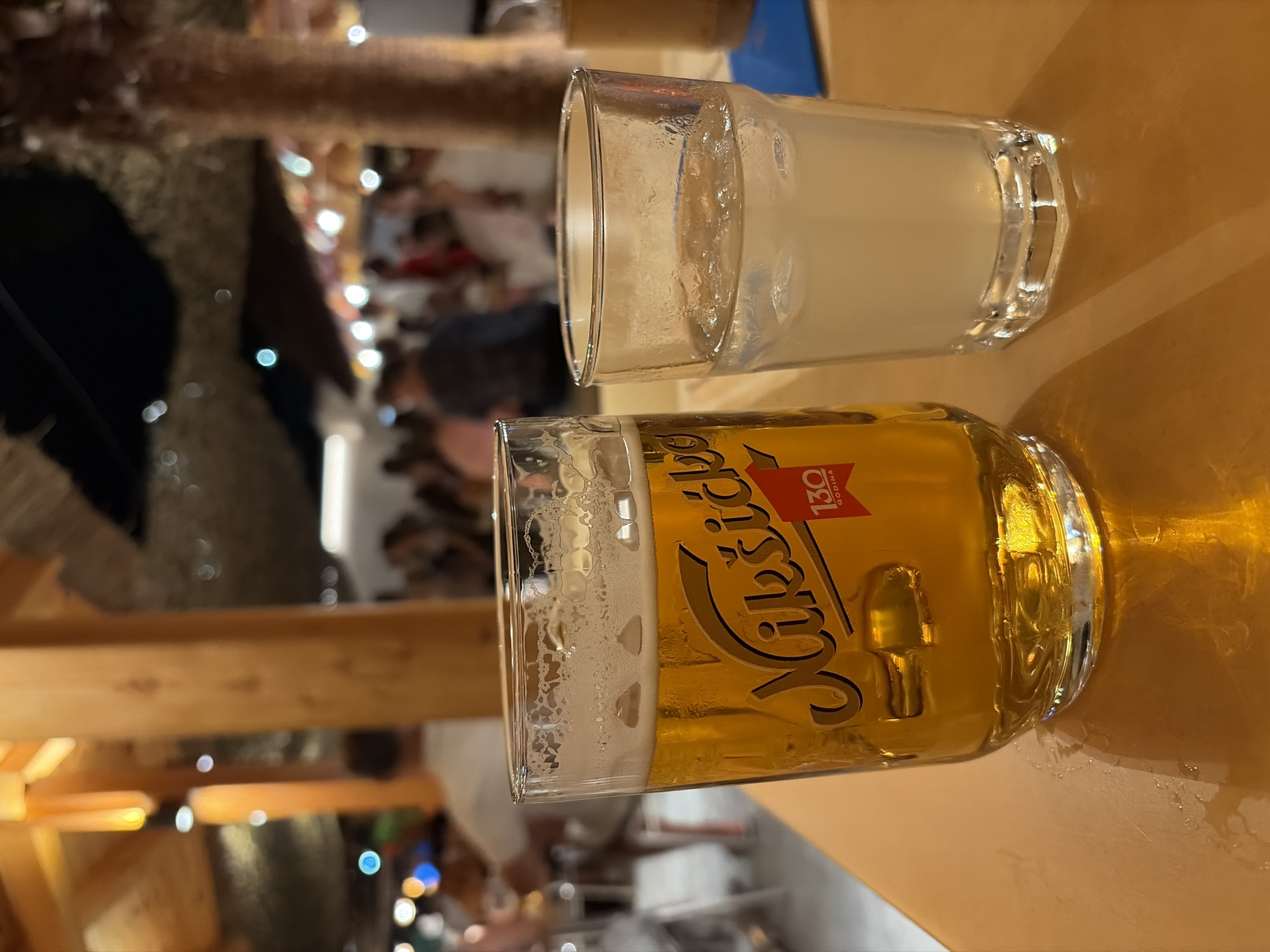 In Boreti, Montenegro, two glasses of beverages are prominently placed on a wooden table. The glass on the left contains a golden-colored liquid, identified by its logo as Nikšićko pivo, a popular Montenegrin beer, celebrating its 130th anniversary. The glass on the right holds a cloudy, light-colored drink with ice. The background is softly blurred, showing a lively outdoor setting with people mingling, illuminated by warm lights, suggesting a restaurant or bar environment with thatched roofing overhead.