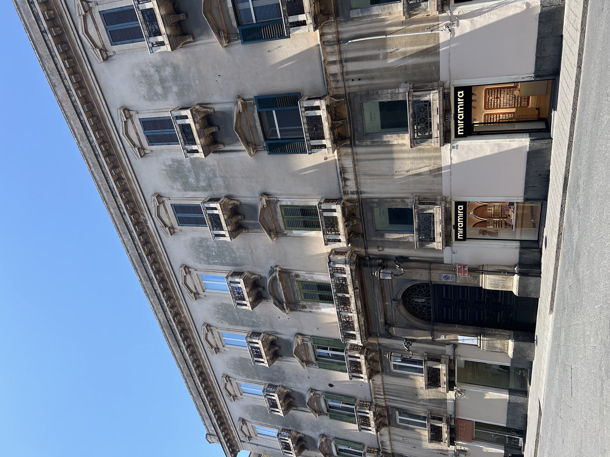 This media file shows the facade of an old building in Catania, Italy. The building has multiple stories with numerous windows, many of which are adorned with small balconies and decorative elements. Some windows have green shutters, while others are open, revealing the interior or reflecting the blue sky. The architecture appears historic, with ornate cornices and detailing along the facade.
On the ground floor, there are storefronts. One storefront clearly displays the name "miramira" in large, illuminated letters, with a display of what appear to be cosmetic or perfume products inside. Another storefront below it also has "miramira" branding, suggesting a retail establishment. Further down, there's another storefront with a sign that partially reads "FREE".
The image is taken from a slightly angled perspective, looking upwards at the building. The bright blue sky suggests it is daytime, and the strong shadows cast by the architectural features and balconies indicate a sunny day, likely mid-morning or early afternoon. The overall scene depicts a street view in a European city, characterized by its classical architecture and commercial activity. There are no people visible in the image. A parking sign and a numbered address sign (111) are visible near the large dark entrance doors.