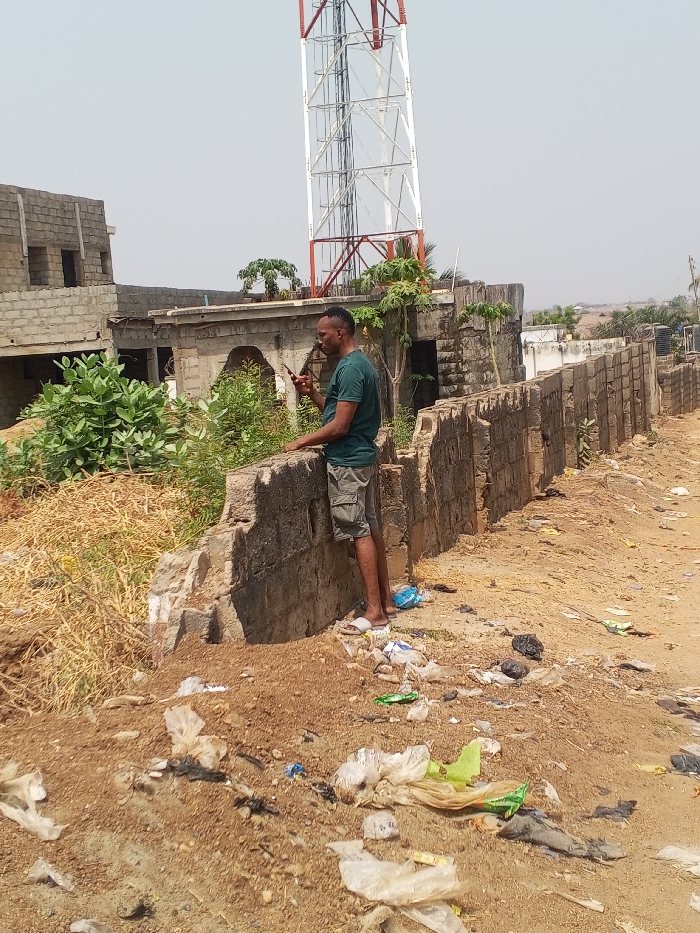 This image captures a daytime scene in Zango-ndaji, Nigeria. A male individual, appearing to be an adult, stands on a dirt path next to a partially dilapidated concrete block wall. He wears a dark green short-sleeved t-shirt, light-colored cargo shorts, and white slide sandals. The man holds a dark-colored smartphone in his right hand, actively looking at its screen, while his left forearm rests on the top of the concrete wall.
The immediate foreground consists of bare reddish-brown earth heavily strewn with litter, including numerous plastic bags, wrappers, and other discarded debris. Behind the man and the wall, dense green shrubs and some dry, yellowish vegetation are visible. In the background to the left, an unfinished two-story building constructed from concrete blocks stands with exposed rebar. To the right of this building, another structure with an arched opening and more vegetation is present. Dominating the upper background is a tall, red and white lattice telecommunication tower against a clear, pale blue sky. Further in the distance, additional buildings and sparse greenery are visible under the bright sky.