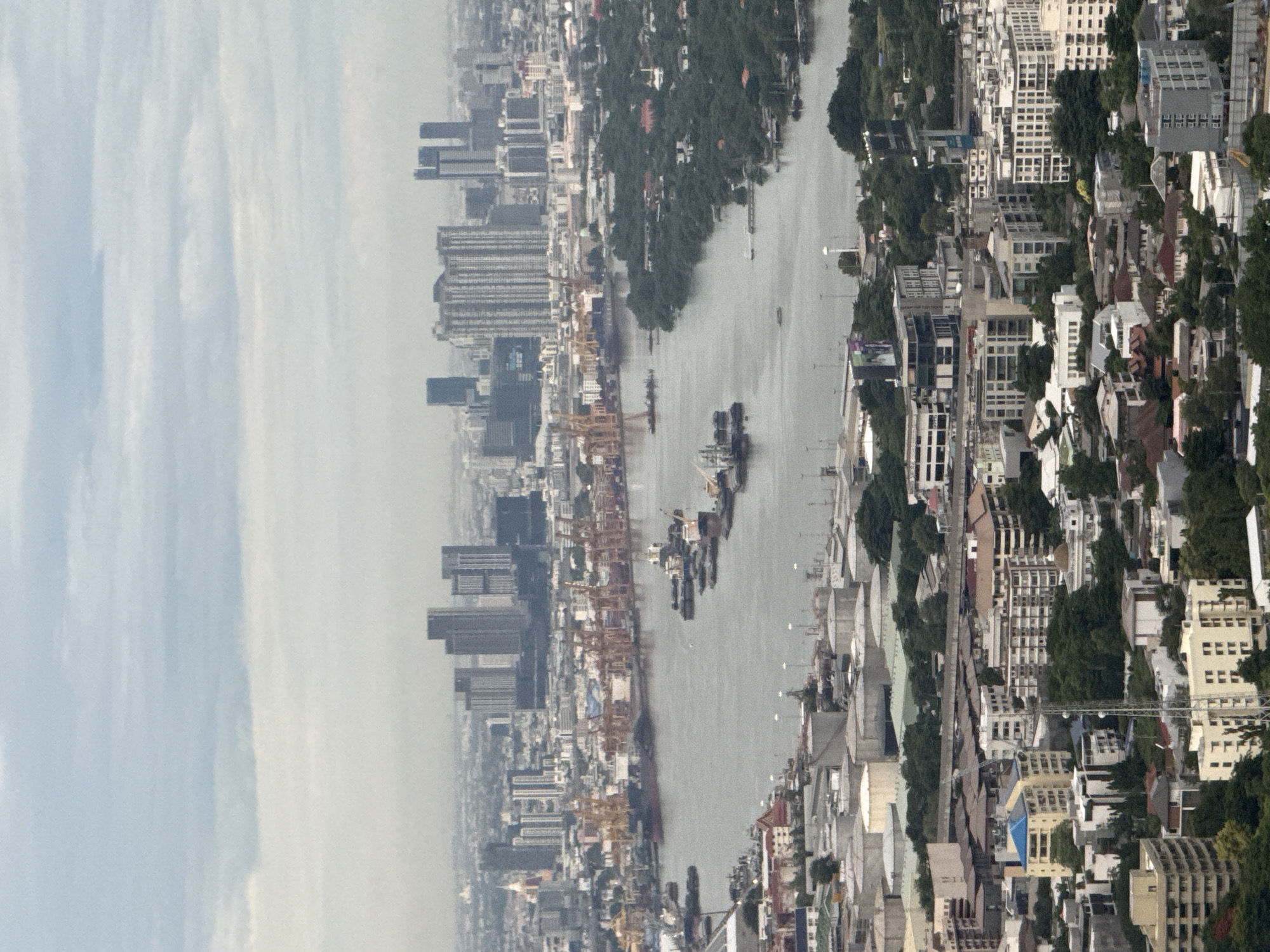 This is an aerial, high-angle, vertical, medium shot photograph of the Chao Phraya River and its surrounding urban landscape in Bangkok, Thailand. The sky is overcast with a light grey cloud cover. The river, occupying the central portion of the image, is wide and reflects the dull sky. A busy port area is visible along the left bank of the river, featuring numerous cranes of varying heights and colors, predominantly orange, and several large cargo ships docked. Further inland on the left, there are industrial storage tanks and complex buildings. The right bank of the river is lined with dense green trees, with some houses and smaller buildings nestled within the foliage. Beyond the river and port, the cityscape of Bangkok unfolds with a dense concentration of buildings, including numerous high-rise apartment buildings and office towers, many of which are modern glass and steel structures. In the foreground, more urban development is visible, with a mix of residential buildings, some appearing to be older low-rise structures and newer apartment blocks. Some industrial facilities with large white cylindrical tanks are also present in the lower left quadrant of the image. There are no visible individuals in the frame. The overall scene depicts a bustling port and a dense, expansive urban environment typical of a major metropolitan city.