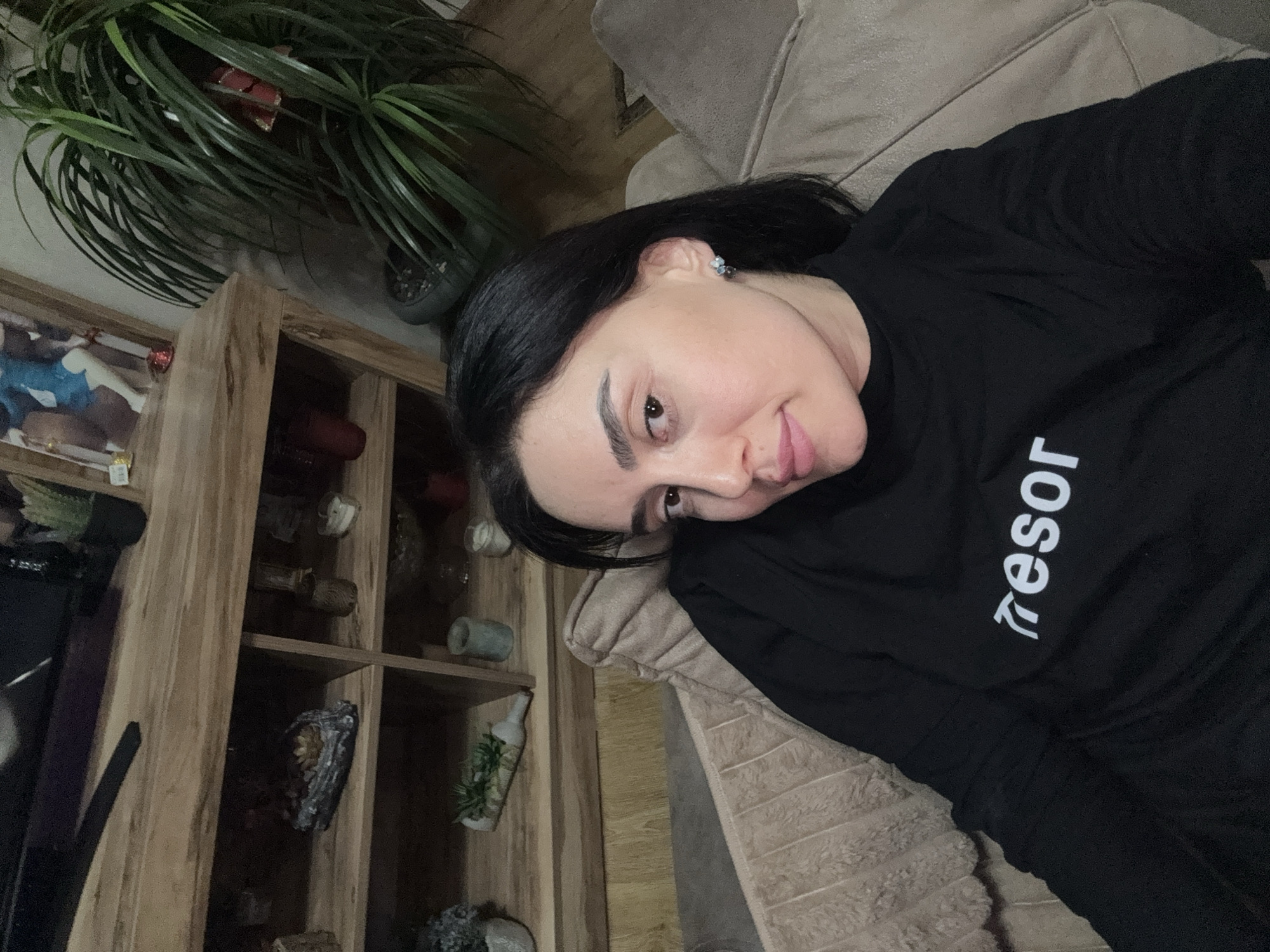 A woman is reclining on a beige sofa in an indoor setting, possibly in a private residence. She is wearing a black long-sleeved shirt with the word "tresor" printed in white on the chest. Behind her is a wooden shelving unit filled with decorative items. To the left of the shelving unit is a potted plant with long, green leaves. The exact location in Tbilisi, Georgia cannot be determined from the image.