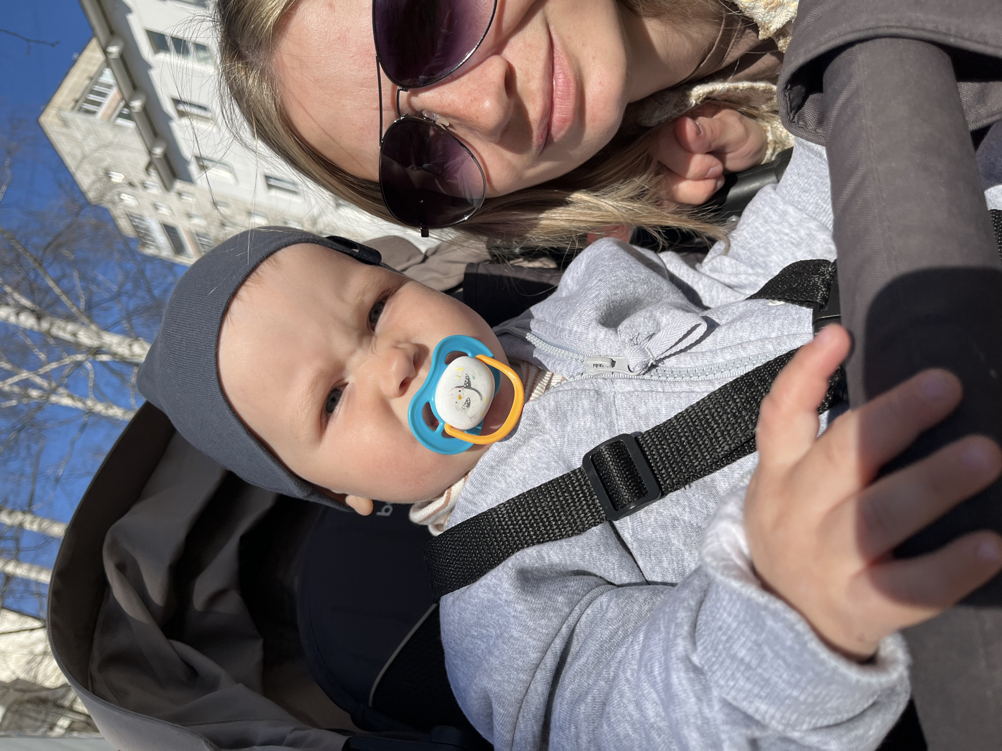 The image shows a baby lying in a stroller in Chaikovsky, Russia, based on the city context. The baby is wearing a grey hat and a grey hooded sweatshirt and has a blue and orange pacifier in its mouth. The baby is secured in the stroller with black straps. A woman, likely the mother, is positioned behind the stroller, partially visible with sunglasses on. A building and trees are visible in the background, indicating an outdoor setting. It appears to be a sunny day.