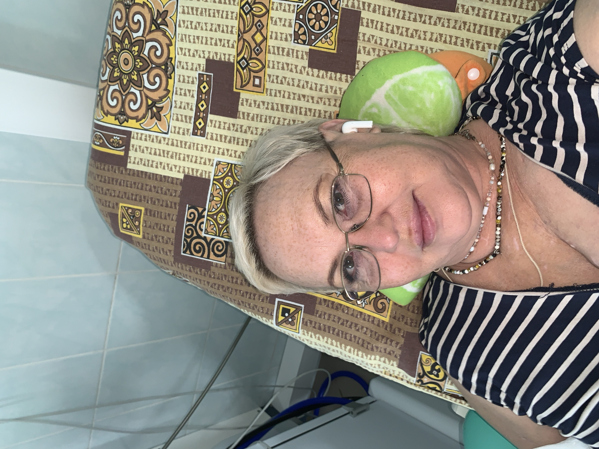An adult female, characterized by short, light-colored hair, freckles, and gold-framed glasses, is depicted in a close-up, upward-facing shot, suggesting a selfie. She is lying on a bed covered with a brown, beige, and yellow patterned mattress or blanket. She wears a dark blue and white striped top, multiple beaded necklaces, and a white wireless earbud in her right ear. Her head rests on a small, green and orange pillow shaped like a vegetable. In the background, to the left, a light blue tiled wall is visible, along with medical or technical equipment featuring white components, blue hoses, and metallic elements. The scene is situated in a medical facility in Chaikovsky, Russia.