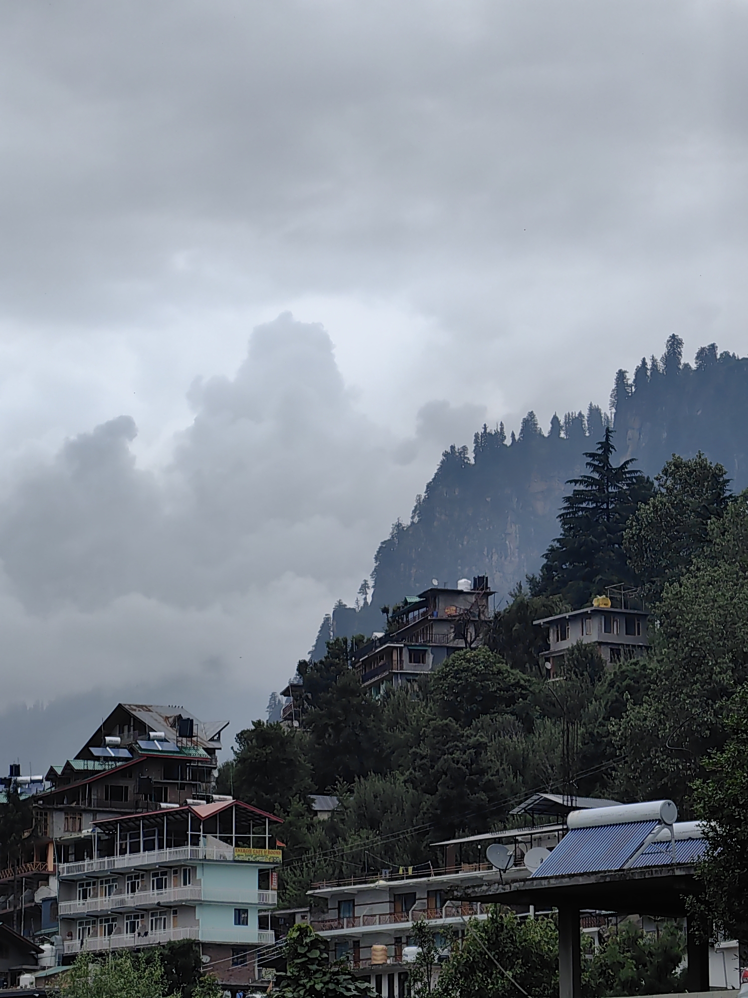 The image depicts a cloudy, overcast day in Vashisht, India. The scene is dominated by a steep, tree-covered mountainside with several buildings constructed into the slopes. The architecture is varied, ranging from multi-story hotels with balconies to smaller, more rustic structures. Many of the buildings have visible solar water heaters on their roofs, suggesting a practical approach to energy in the region.
The vegetation is lush and green, consisting of a mix of coniferous and deciduous trees, which create a dense forest covering the mountain. The sky is filled with heavy grey clouds, indicating a possibility of rain or recent rainfall. The overall atmosphere is somber and somewhat dramatic, with the mist or low-lying clouds adding a sense of depth and mystery to the mountainous landscape.
There is no visible activity of people in the image, and the focus is on the built environment nestled within the natural landscape. The time of day is difficult to ascertain precisely due to the overcast conditions, but the diffused light suggests it is daytime. The location cues, such as the style of buildings and the mountainous terrain, are consistent with Vashisht, India. One building has text that reads "ENLIGHT CAFE ROOMS".