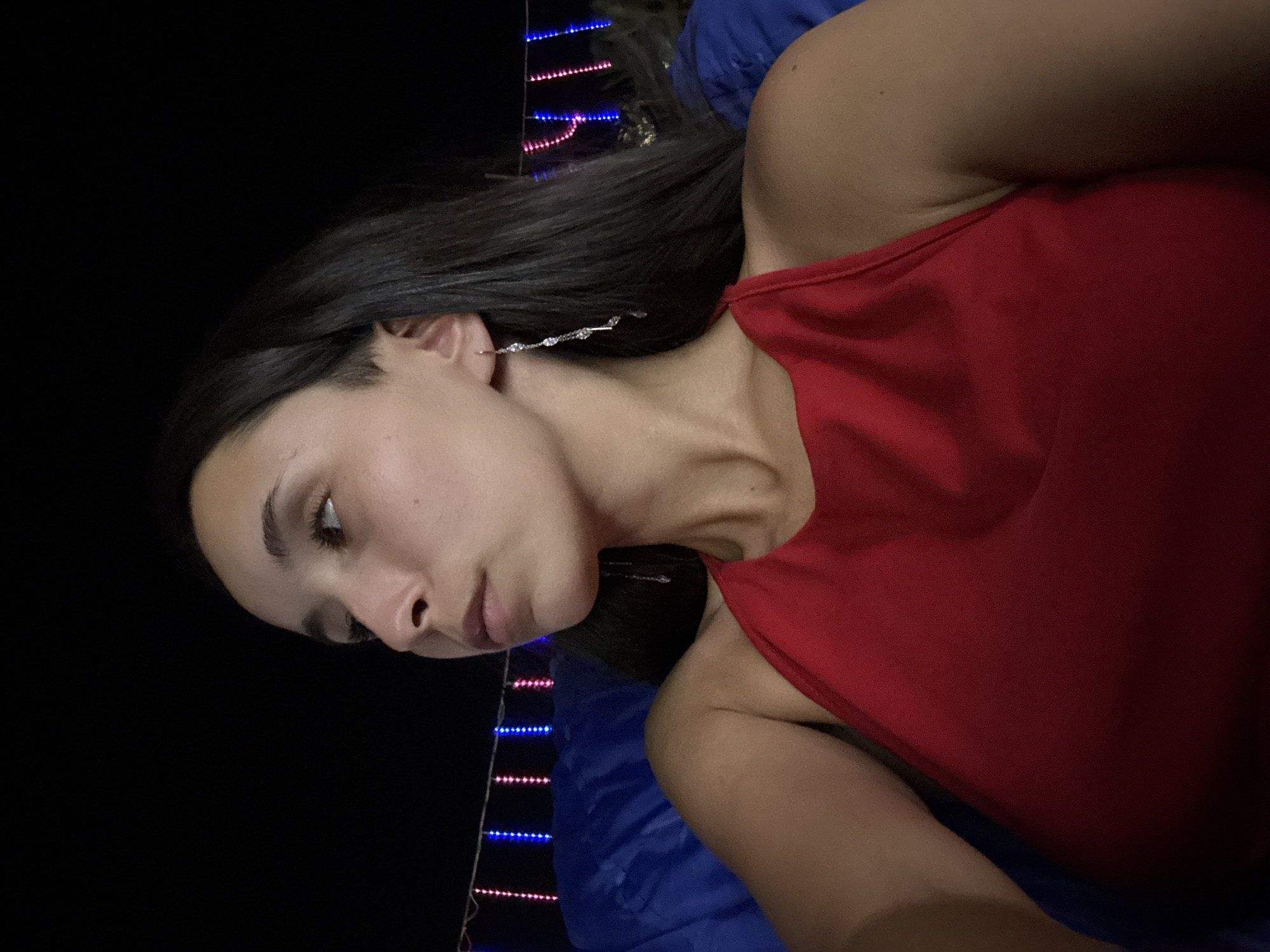 A young woman with dark hair and light skin is depicted in a medium close-up, looking to her left. She is wearing a red halter top and a long, dangling earring on her right ear. The background is dark with a string of red and blue lights visible behind her. She appears to be outdoors at night, possibly in a relaxed or celebratory setting. The blue object behind her could be a cushion or a blanket. The scene is lit with artificial light, likely from the string lights. No specific location in a city is identifiable.