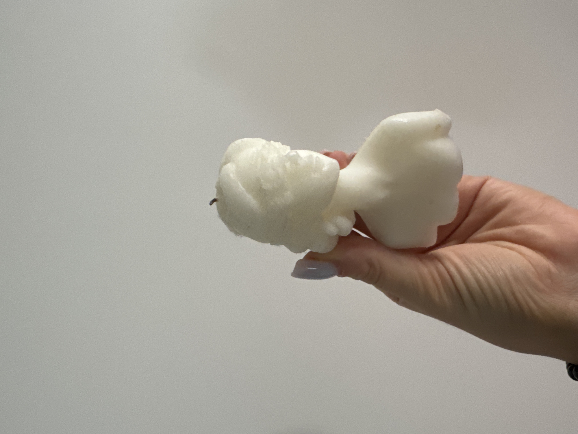 A person's hand, with light skin and manicured nails painted a pale blue, is holding a white candle shaped like a bust of a person. The candle is a pale white and has a wick sticking out of the top. The background is a plain, light gray wall. The image does not contain any information about the location in the city of Murcia, Spain.