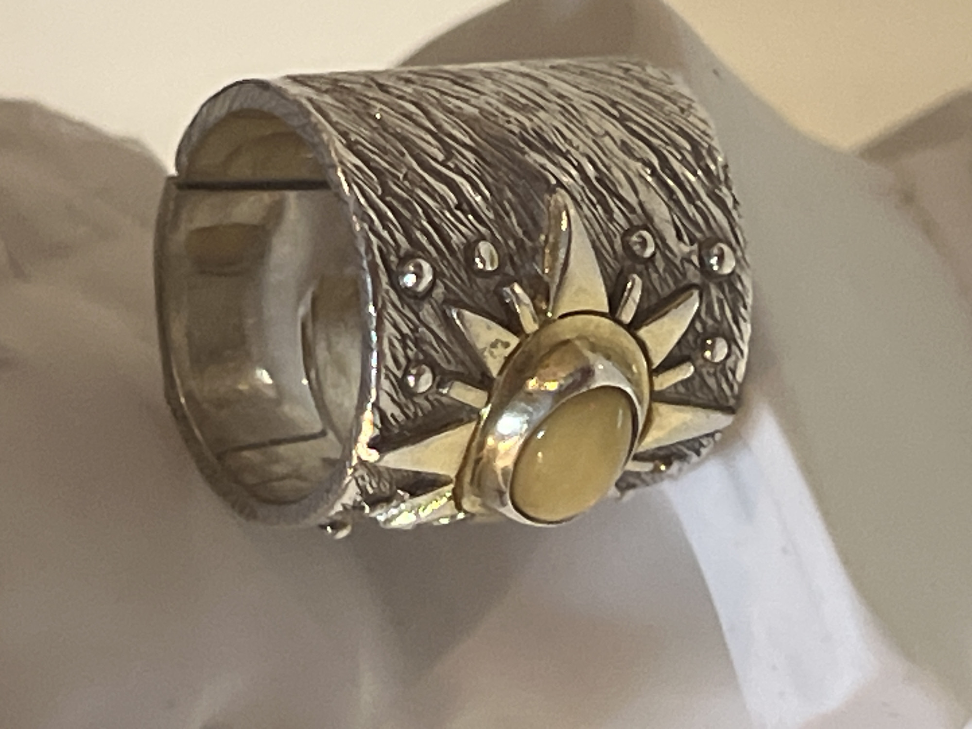 A close-up, slightly high-angle shot shows a wide, textured silver ring with a raised sunburst design. The sunburst is outlined in gold and features a smooth, oval, light yellow or amber stone at its center. The band of the ring has a rough, linear texture reminiscent of wood grain and is adorned with small, raised silver dots around the sunburst motif. The ring is positioned against a blurred, light-colored background, with a shadow cast on the right side. The ring appears to be resting on a white surface, possibly a bust or display. No people are visible in the image.