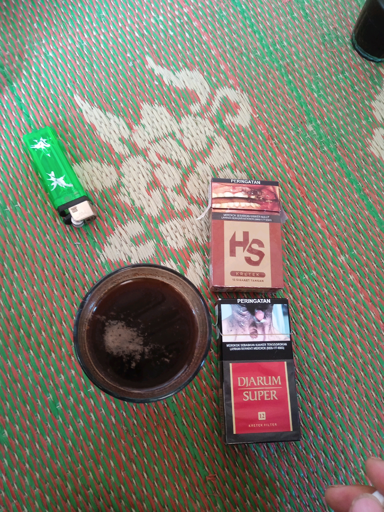 A top-down view captures a collection of items on a woven mat. In the upper left corner, a green lighter with white star-like designs is positioned diagonally. To the right and slightly below the lighter, a dark-colored glass filled with a dark liquid, presumably coffee, sits at an angle. Two packs of cigarettes are placed side-by-side below the glass. The top cigarette pack is predominantly brown and orange with "HS" prominently displayed, and smaller text indicating "KRETEK" and "12 SIGARET TANGAN". The bottom cigarette pack is black and red with "DJARUM SUPER" in large white letters, a "12" in a red box, and "KRETEK FILTER" below. Both cigarette packs feature "PERINGATAN" (Warning) at the top, followed by graphic images related to health warnings. The woven mat has a pattern of green, red, and off-white diagonal lines. The scene appears to be outdoors, possibly in a rural or semi-urban setting in Kelurahan, Indonesia, given the commonality of such mats and cigarette brands in the region. No individuals are visible in the frame.